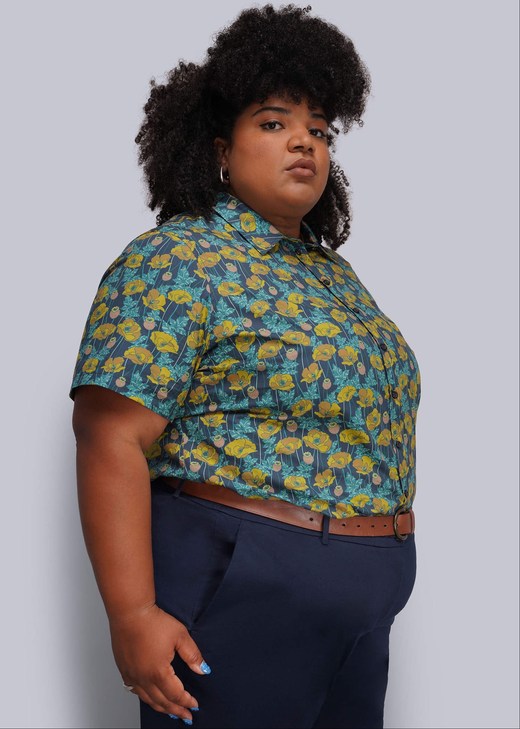 1X=A plus-size model wearing a short-sleeve button-up shirt with a blue, yellow, and green floral pattern, paired with navy blue trousers and a brown belt, against a solid light gray background.