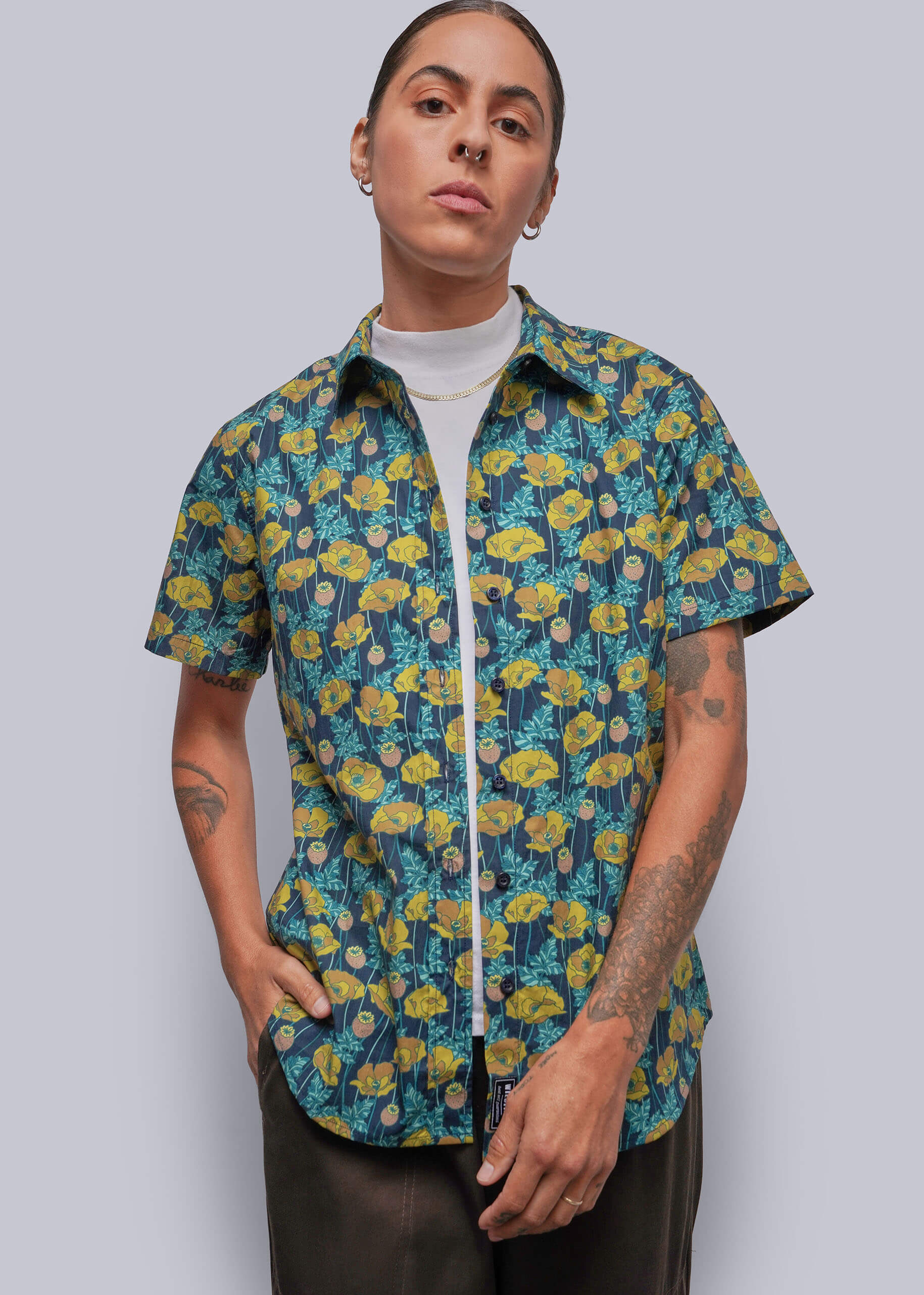 S=A model wearing a short-sleeve button-up shirt with a vibrant yellow and teal floral pattern, layered over a white t-shirt and dark brown pants, set against a solid gray studio background.