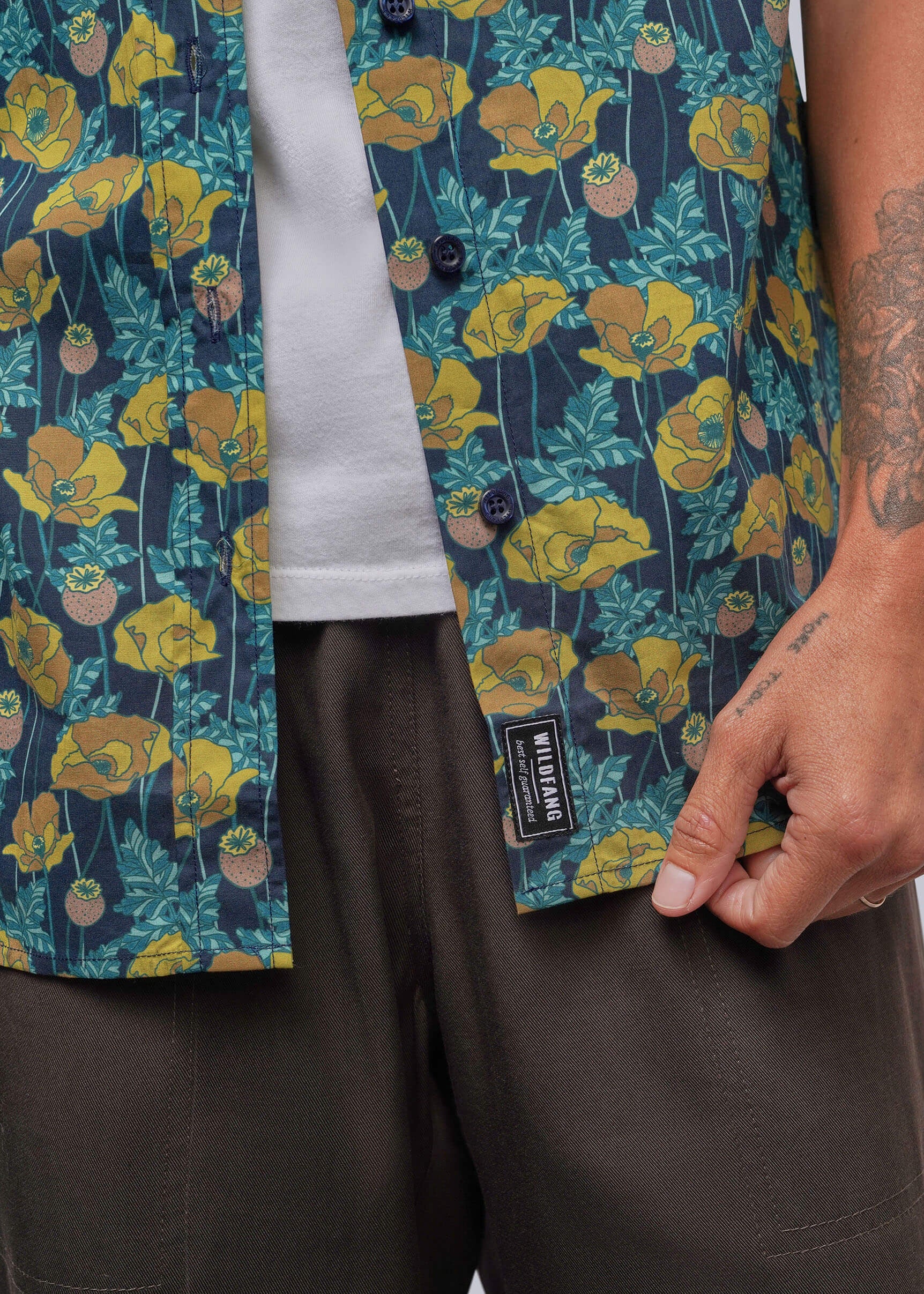 S=Close-up of a person wearing a short-sleeve button-front shirt with a dark teal background and a yellow floral print, layered over a white t-shirt and paired with dark brown pants. A black brand label reading 'SILKSTITCH' is visible near the hem.