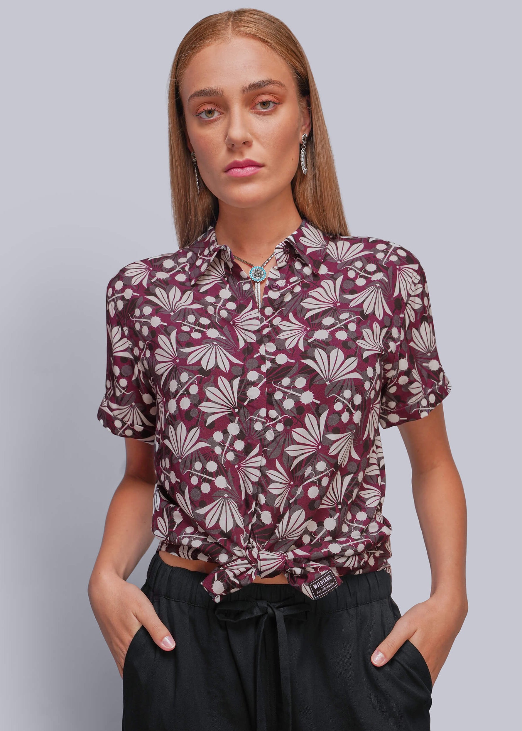 A woman modeling a short-sleeve button-up shirt with a burgundy and beige floral pattern, tied at the waist, paired with black drawstring pants against a solid gray background.