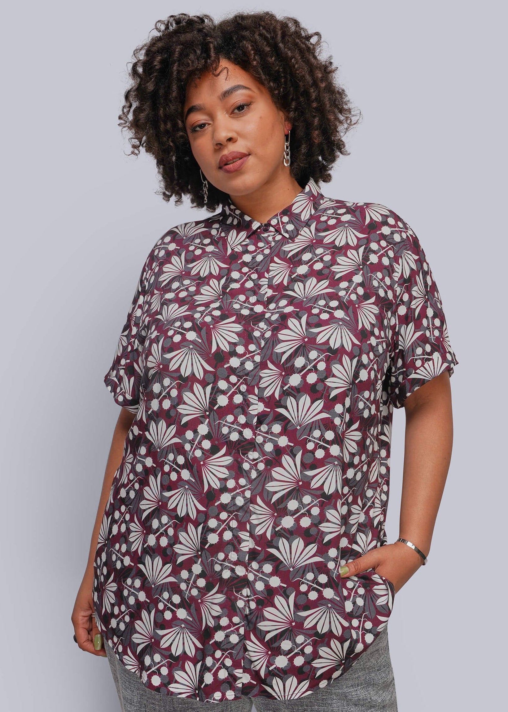 1X=A woman with curly dark hair models a short-sleeve button-up shirt featuring a burgundy, white, and gray floral pattern, paired with gray pants, against a solid light gray background.