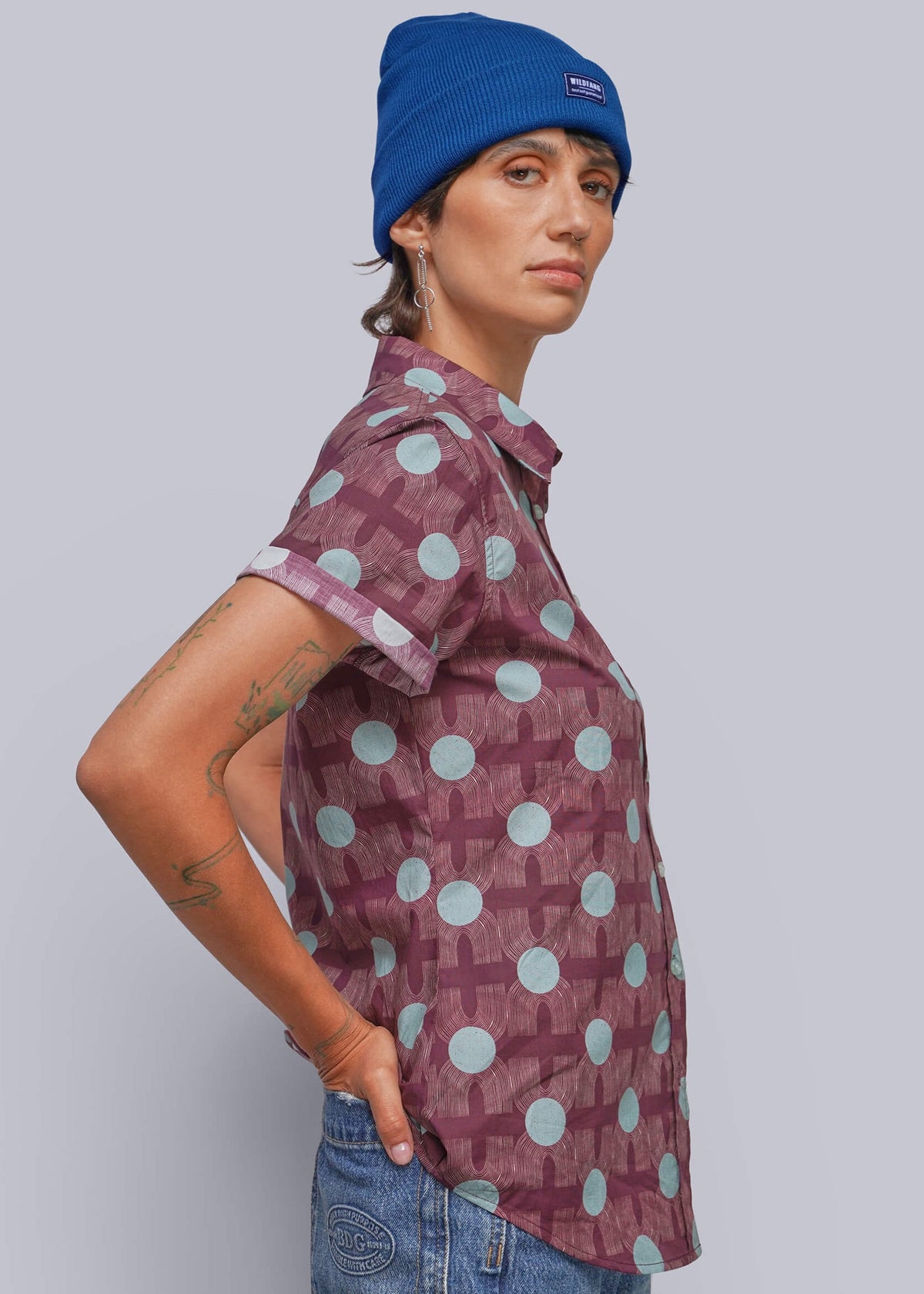 S=A person wearing a blue beanie, a patterned short-sleeve button-up shirt, and blue jeans, shown in profile against a light gray studio background.