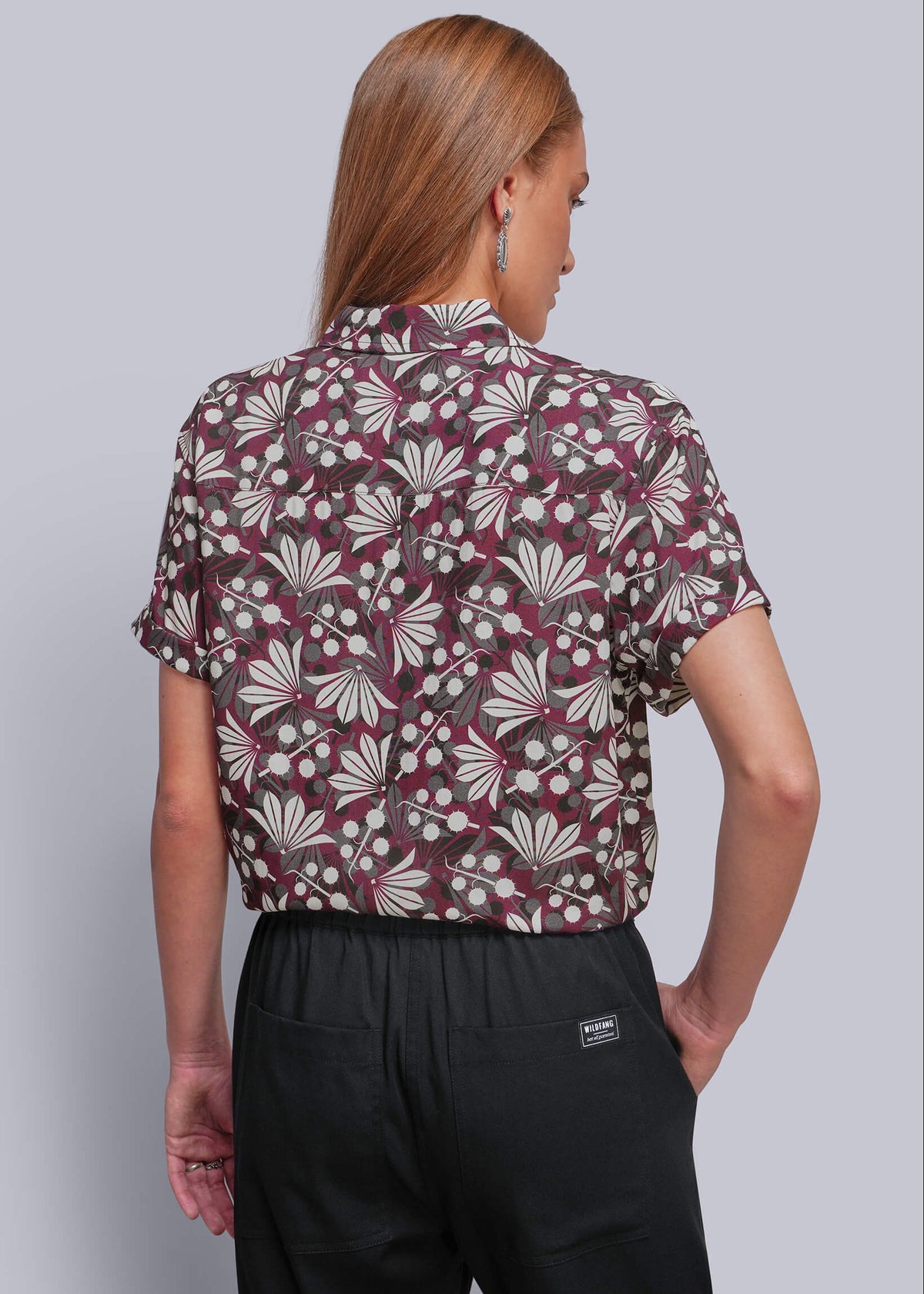 S=A woman shown from the back wearing a short-sleeve button-up shirt with a burgundy, beige, and white floral pattern, paired with black trousers, against a solid gray studio background.