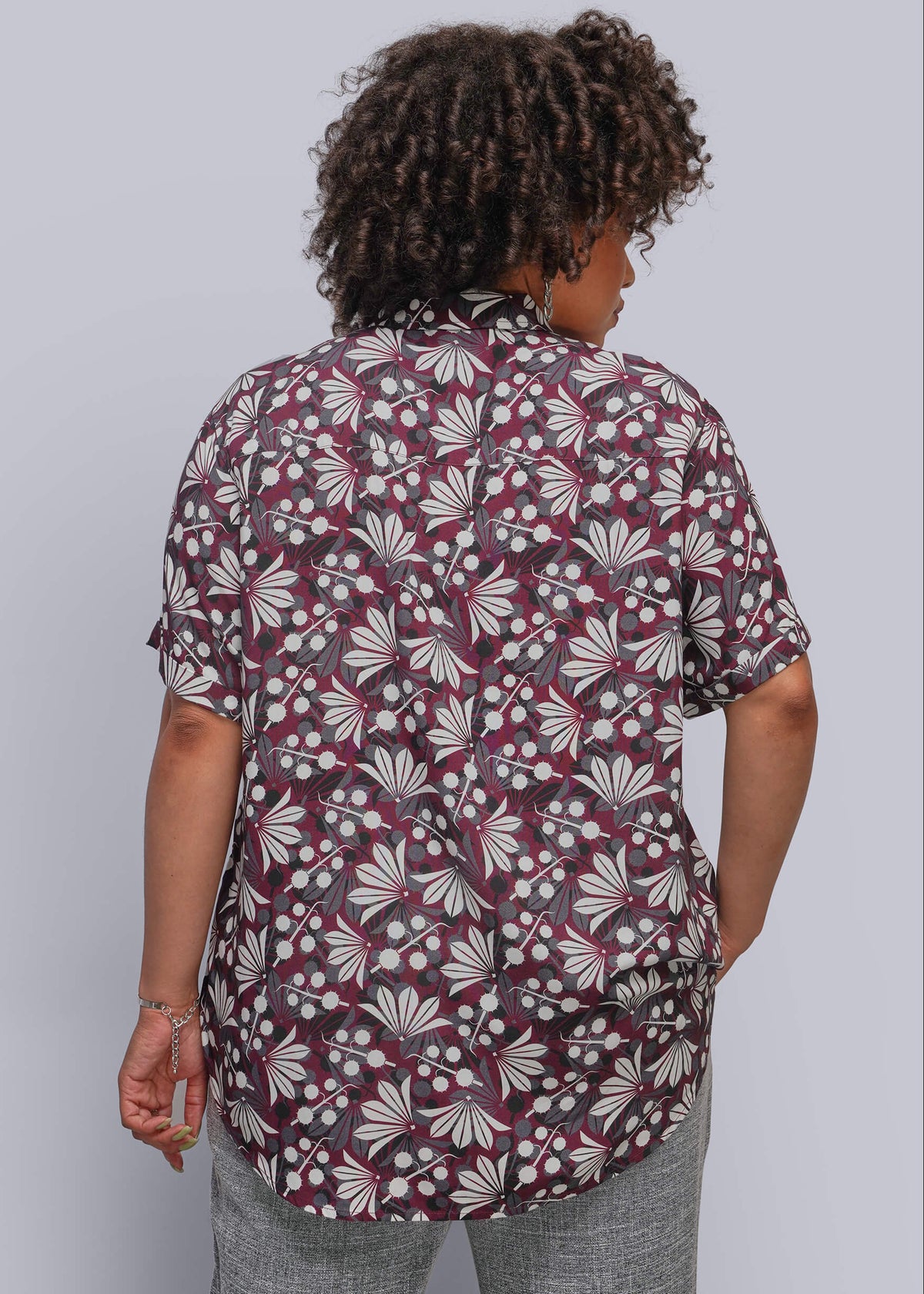 1X=A woman viewed from behind wearing a short-sleeve button-up shirt with a dark burgundy and gray floral pattern, paired with gray pants, against a solid light gray background.
