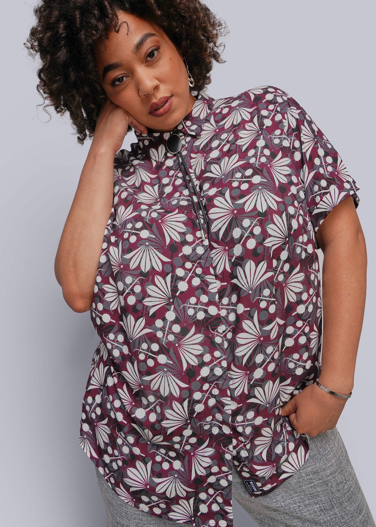 1X=A woman wearing a short-sleeve button-up blouse with a collar and a dark gray, burgundy, and white abstract floral pattern, paired with gray pants, against a solid light gray background.