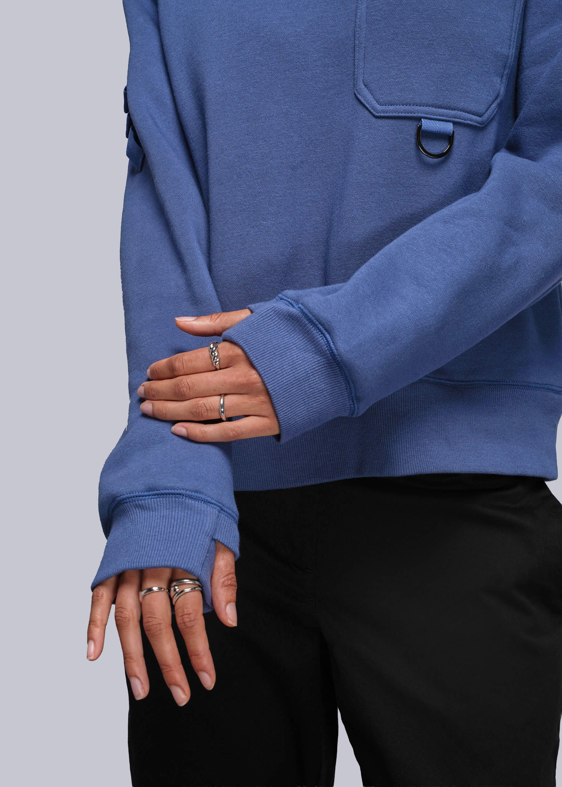 S=Person wearing a blue sweatshirt with a pocket, holding their hand with rings on a light gray background