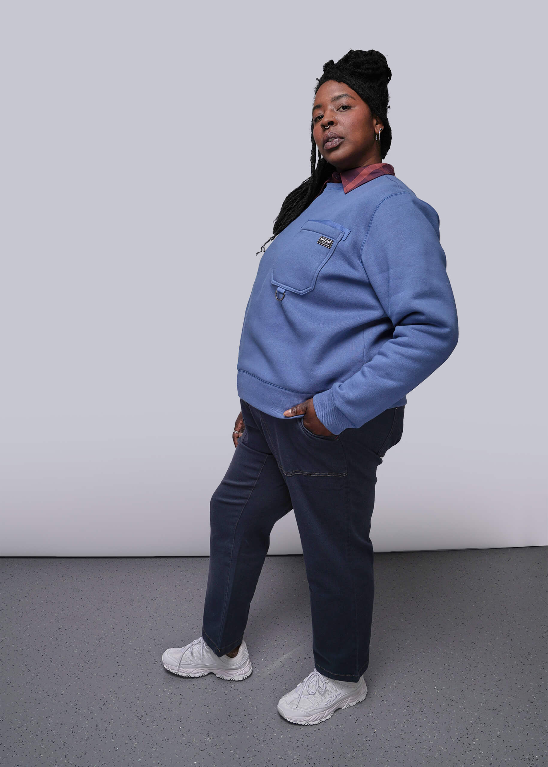 1X=Person wearing a blue sweatshirt and dark pants against a plain background