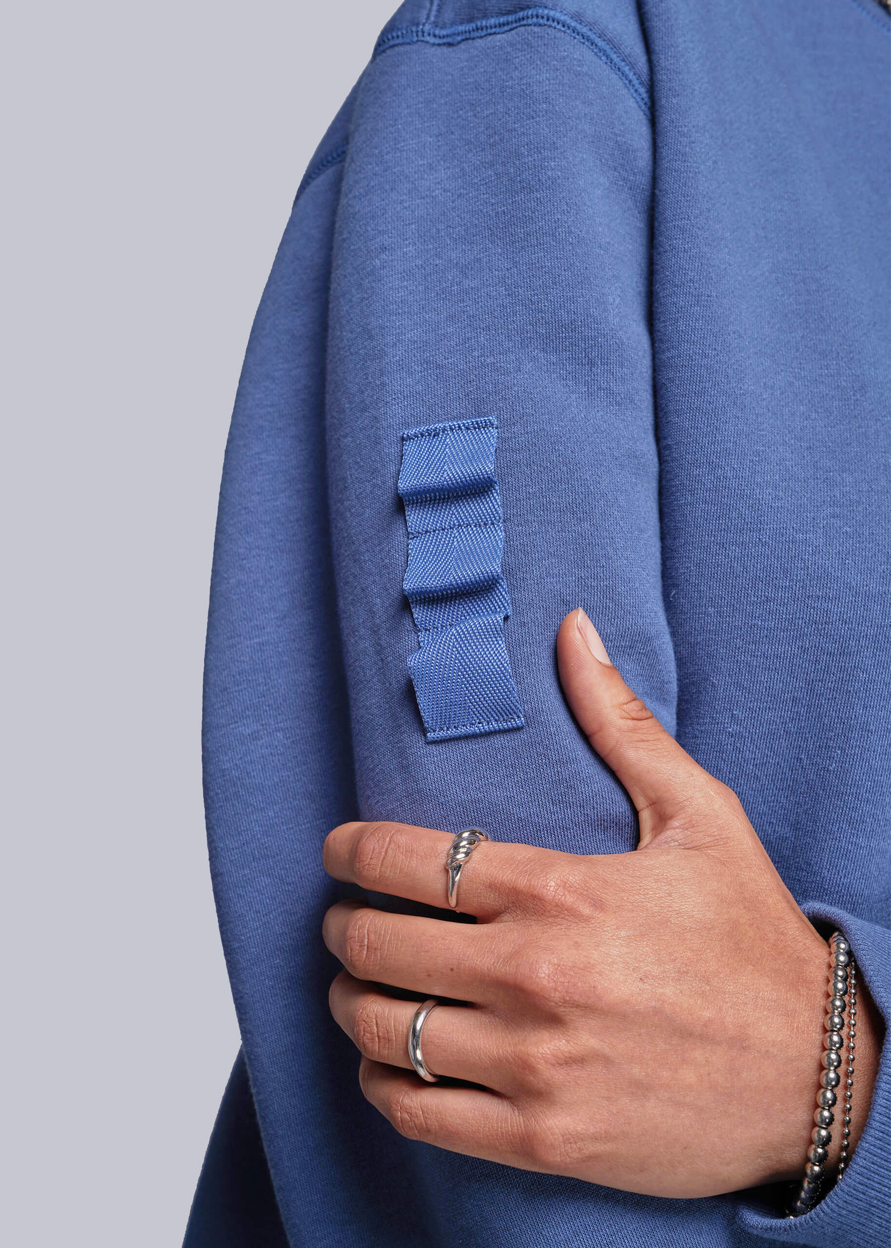 S=Blue sweatshirt with a patch on the sleeve and a hand touching it against a light gray background