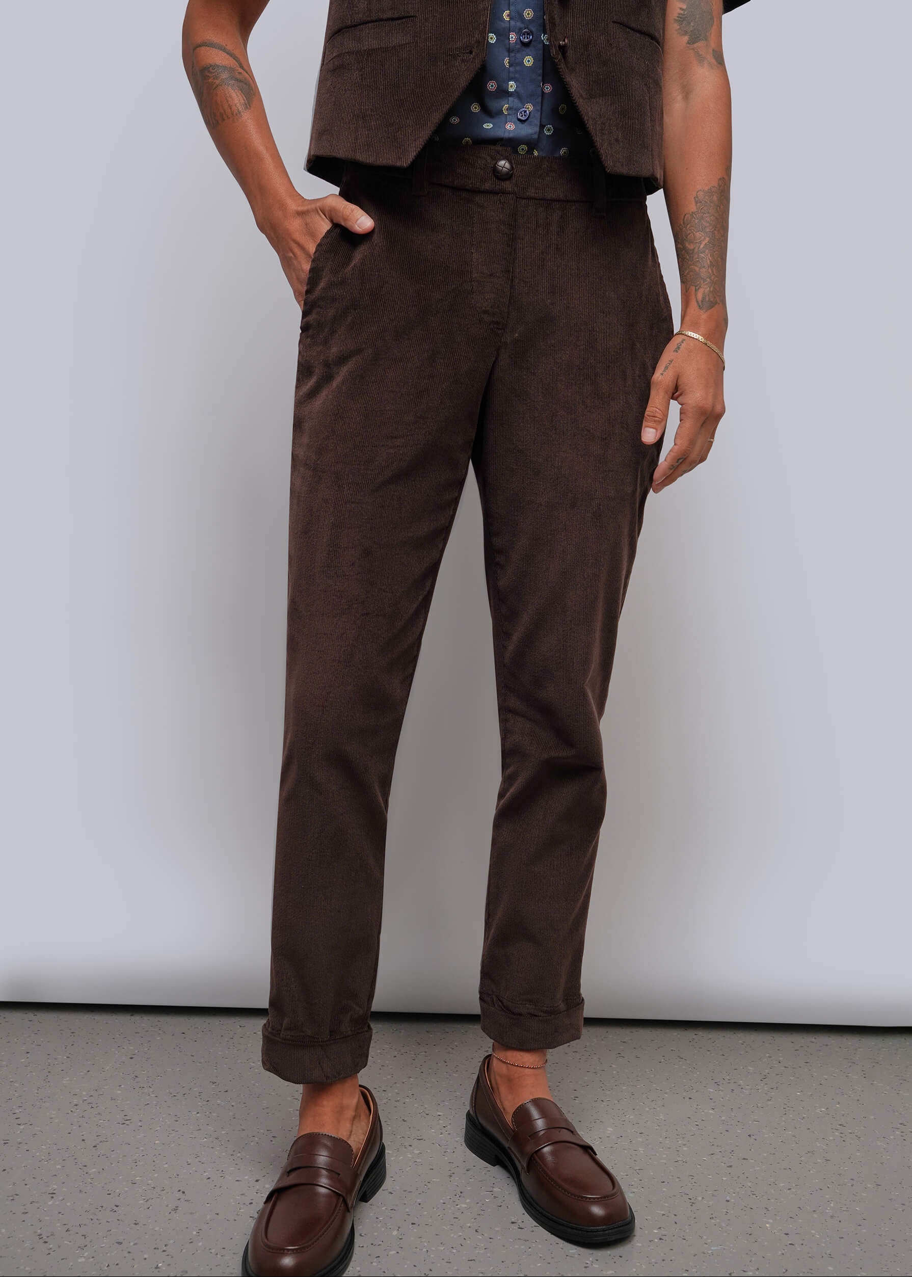 A person wearing dark brown corduroy trousers, styled with a matching brown vest and brown leather loafers against a plain light gray studio background.
