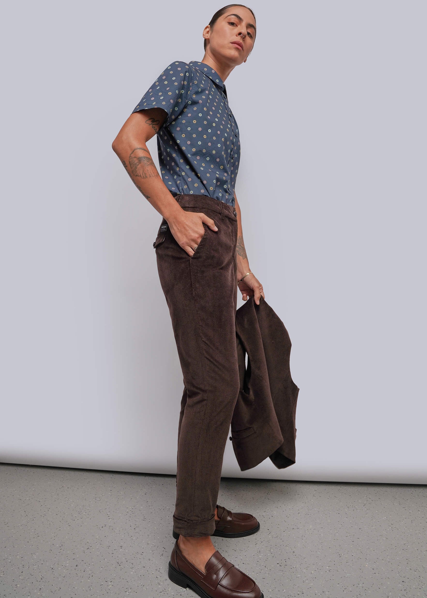 S=A model stands in a studio wearing a blue patterned short-sleeve button-up shirt, brown corduroy trousers, and brown leather loafers, while holding a dark brown jacket.