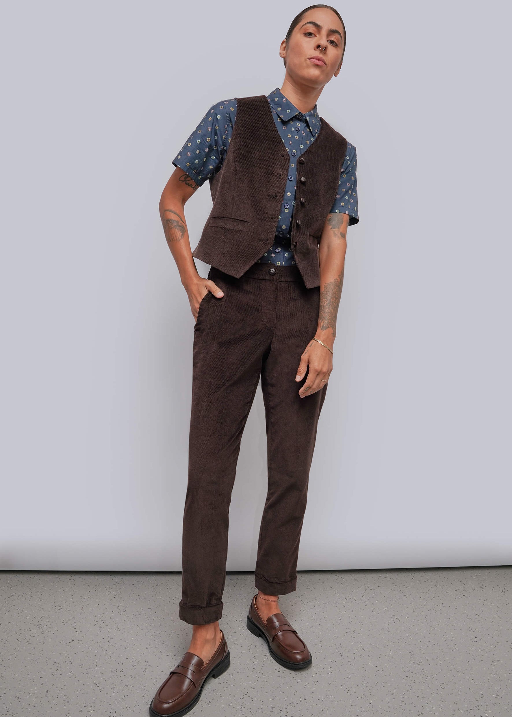 S=A model wearing a dark brown corduroy vest and matching trousers, styled with a blue patterned short-sleeve shirt and brown leather loafers, standing against a light gray studio background.