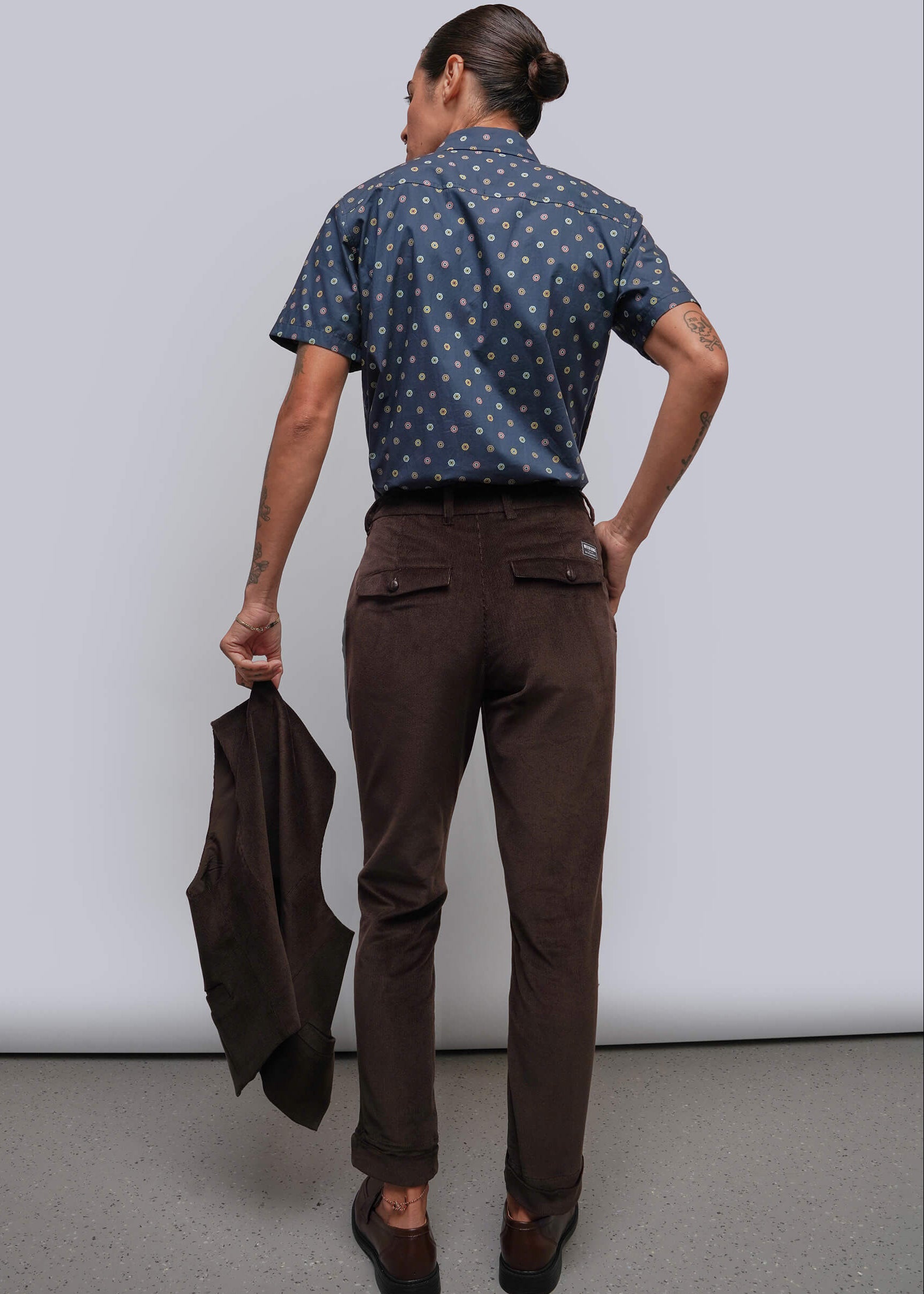 S=A model shown from the back wearing a navy blue short-sleeve button-down shirt, dark brown trousers, and holding a dark brown blazer against a light gray studio background.