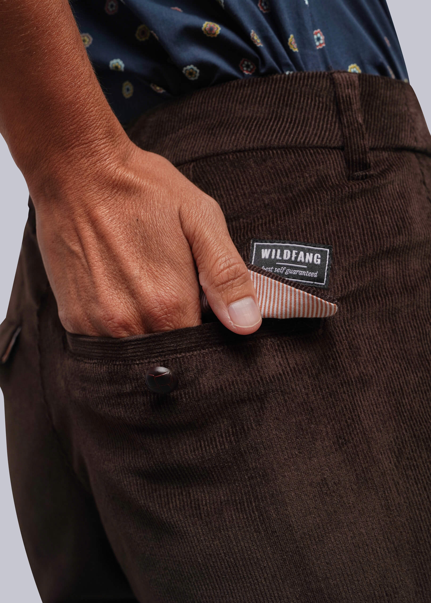 S=Close-up of a person wearing dark brown corduroy trousers, with a hand reaching into the back pocket to pull out a striped paper pocket square. The brand label 'WILDFANG' is visible on the back waistband.