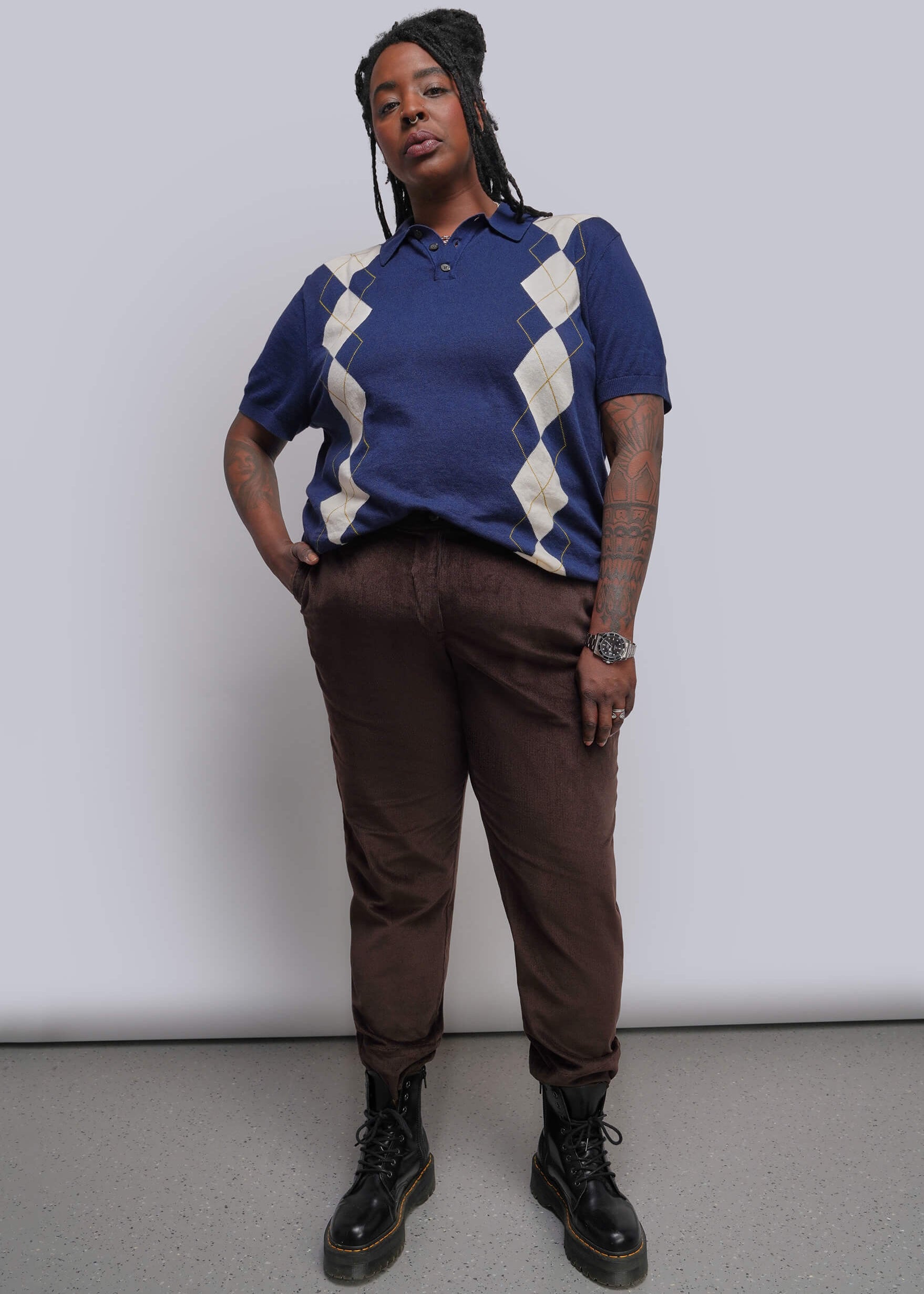 1X=A person modeling a short-sleeve knit polo shirt with a blue base and cream argyle pattern, paired with brown trousers and black lace-up boots, standing against a plain light gray studio background.