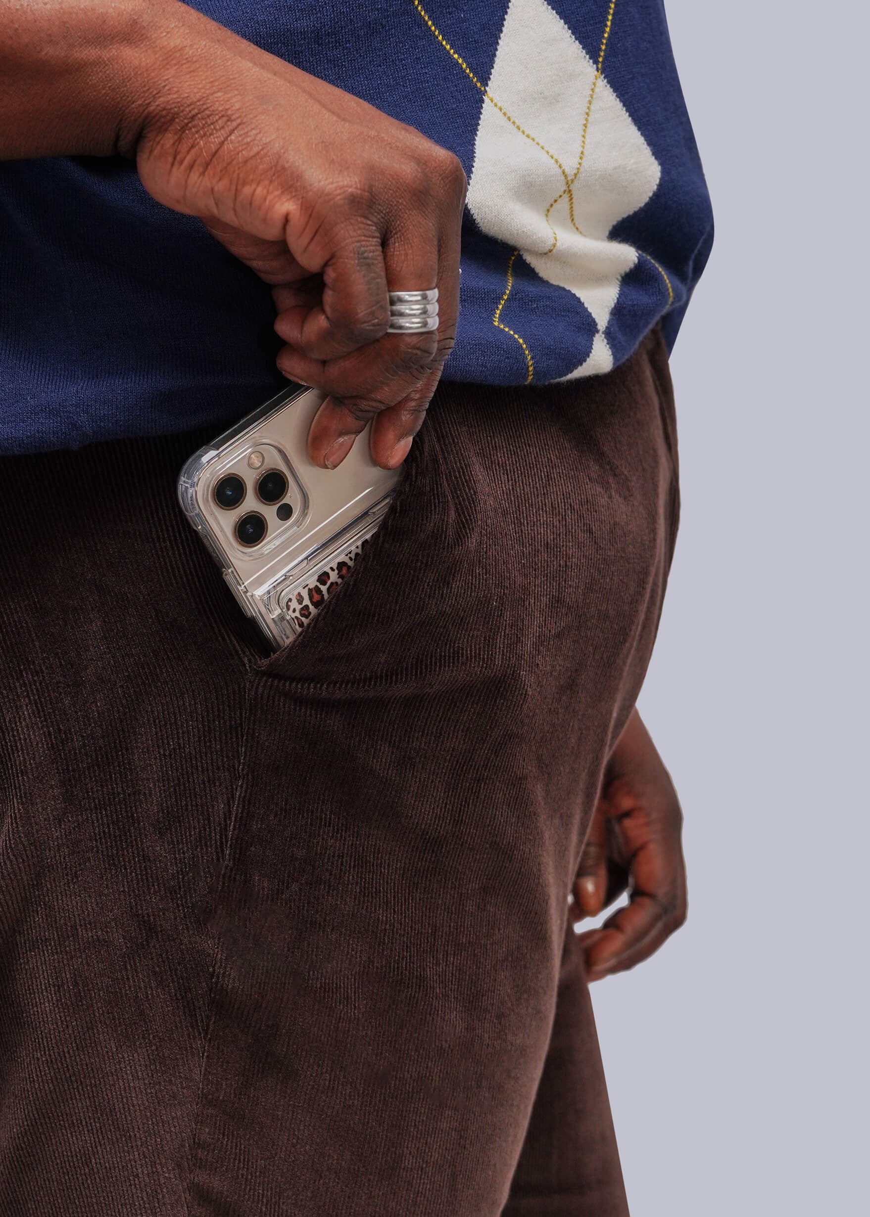 1X=A close-up lifestyle shot of a person wearing brown corduroy trousers and a blue and white sweater, showing a hand reaching into the pocket of the trousers to retrieve a silver smartphone.