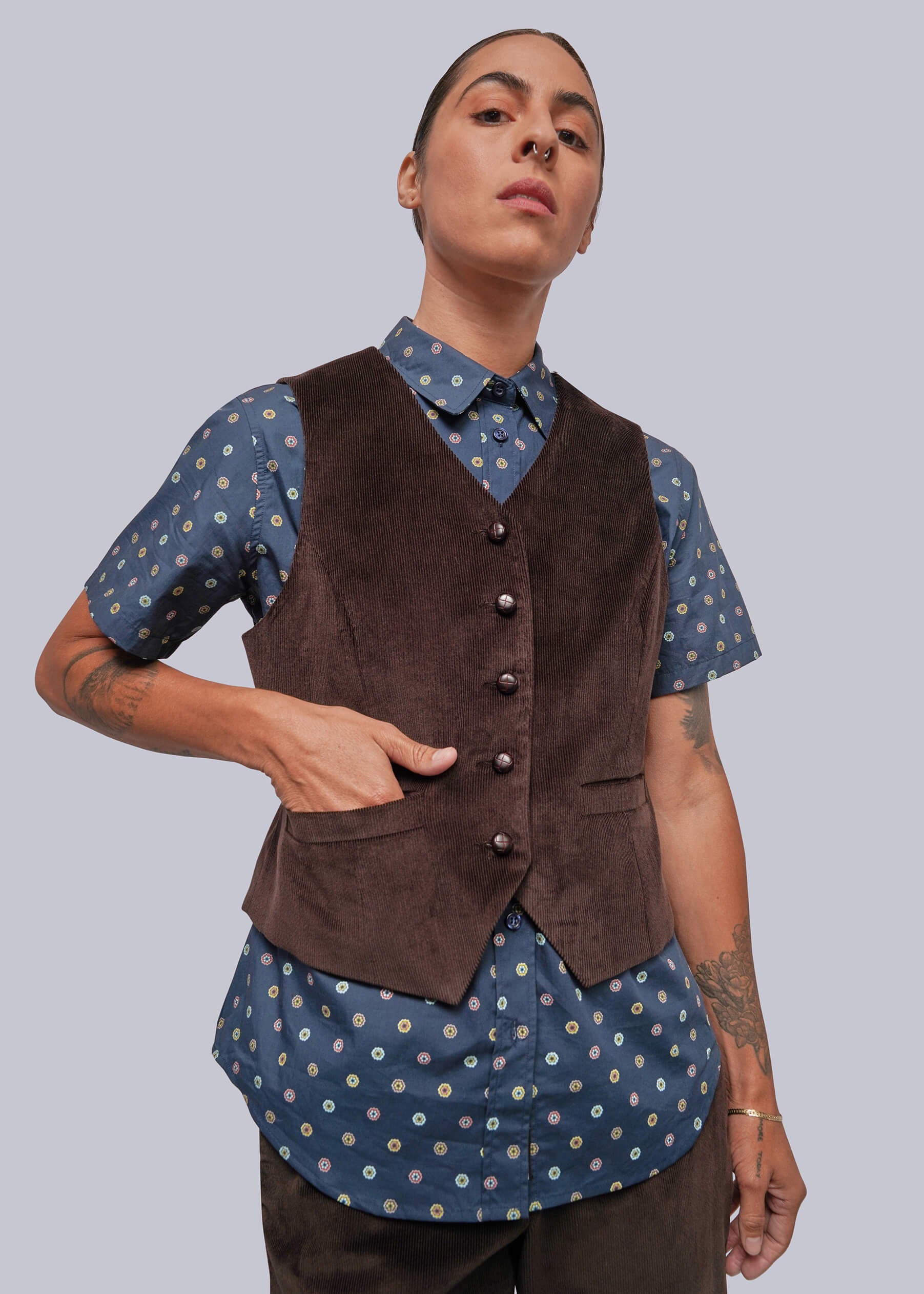 S=A model wearing a dark brown corduroy vest over a blue and white patterned short-sleeve button-up shirt, paired with dark brown corduroy trousers, against a solid gray background.