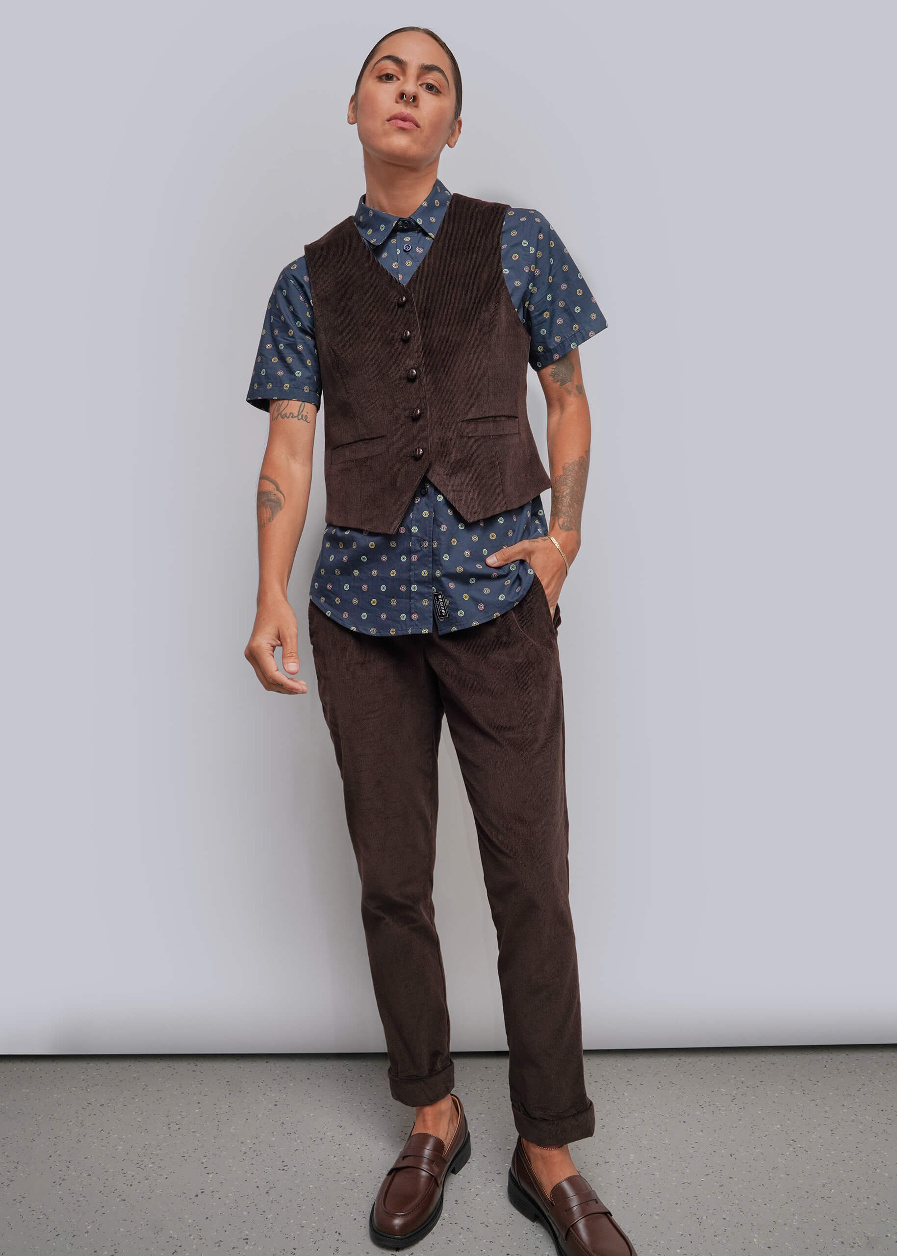 A model wearing a dark brown corduroy vest over a blue patterned short-sleeve button-up shirt, paired with matching dark brown corduroy trousers and brown leather loafers, standing against a light gray studio background.