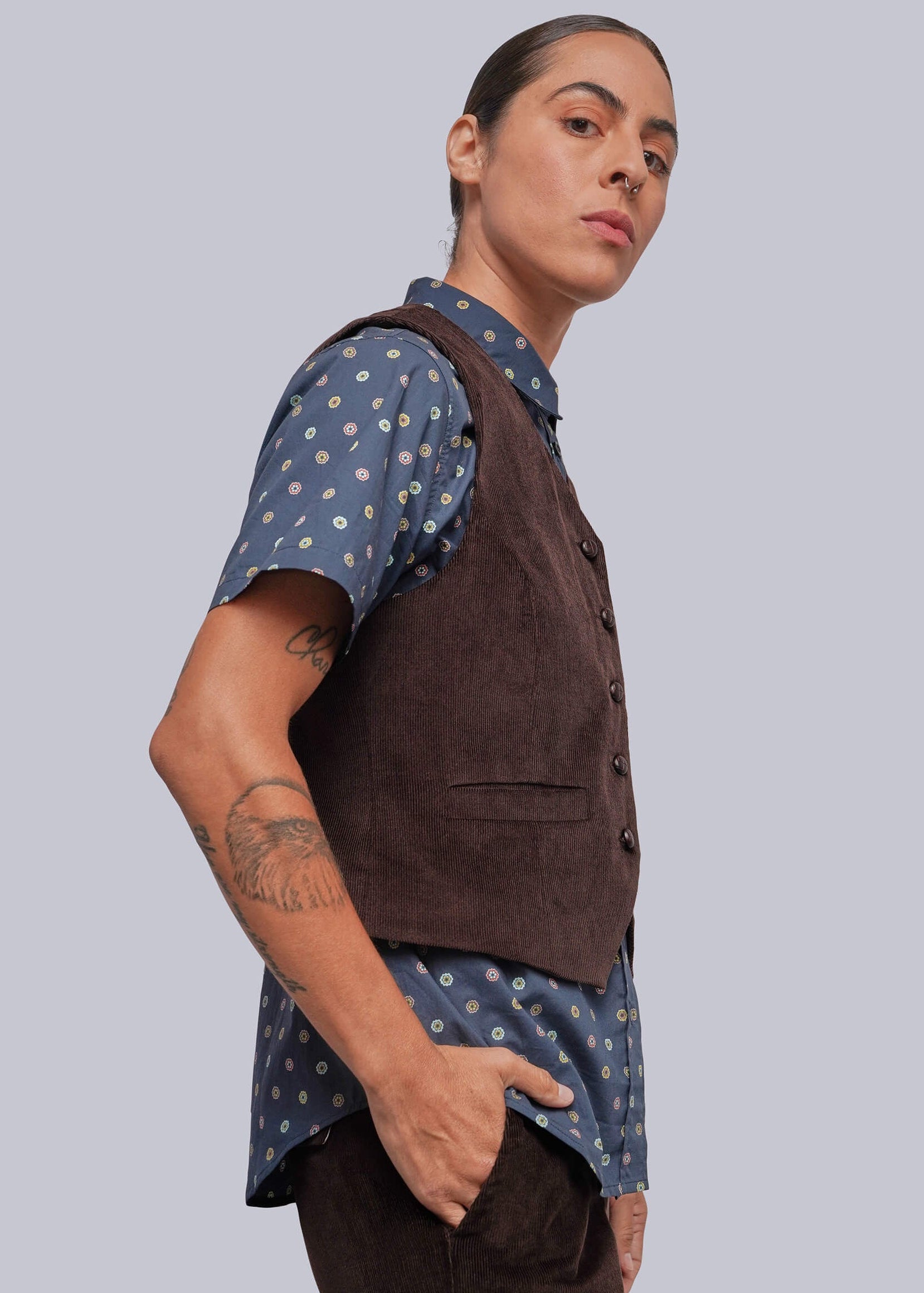 S=A model wearing a dark brown corduroy vest over a blue short-sleeve button-up shirt with a white polka dot pattern, paired with dark brown trousers, set against a solid light gray background.
