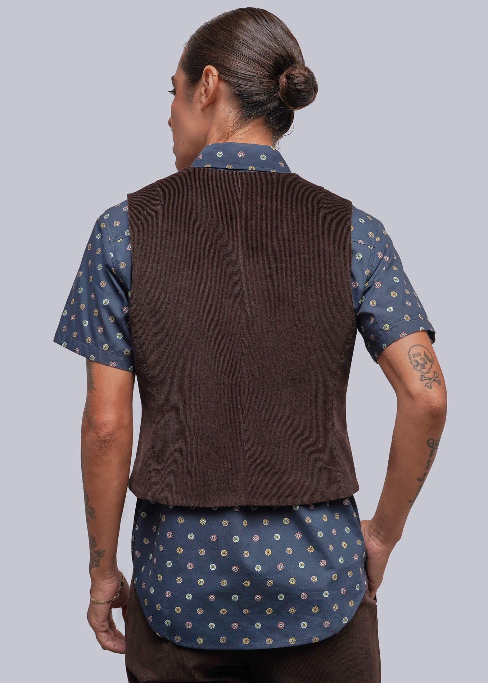 S=A model shown from the back wearing a dark brown corduroy vest over a blue short-sleeve button-up shirt with a white floral pattern, paired with dark brown trousers, against a solid gray background.