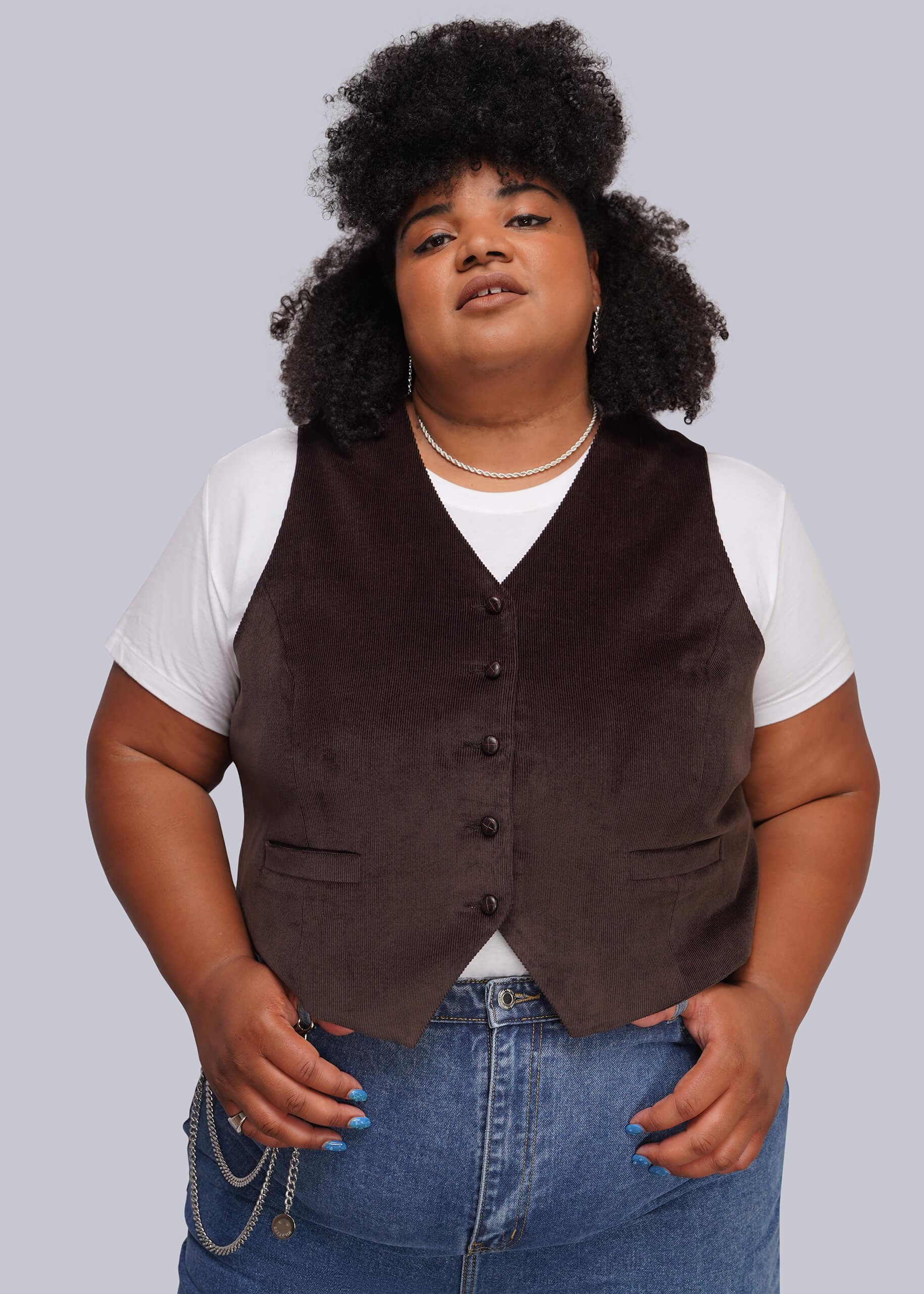 1X=A plus-size model wearing a dark brown corduroy vest over a white t-shirt and blue jeans, standing against a solid gray background.