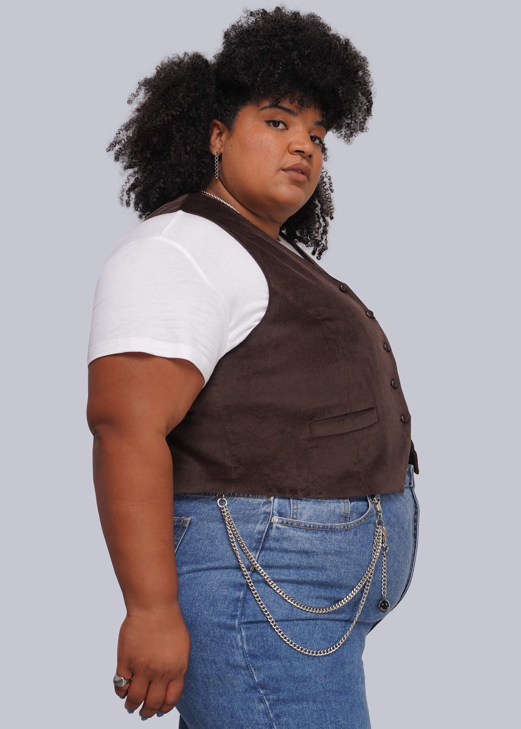 1X=A plus-size model wearing a dark brown corduroy vest over a white t-shirt and blue jeans with silver wallwt chain, set against a solid gray background.