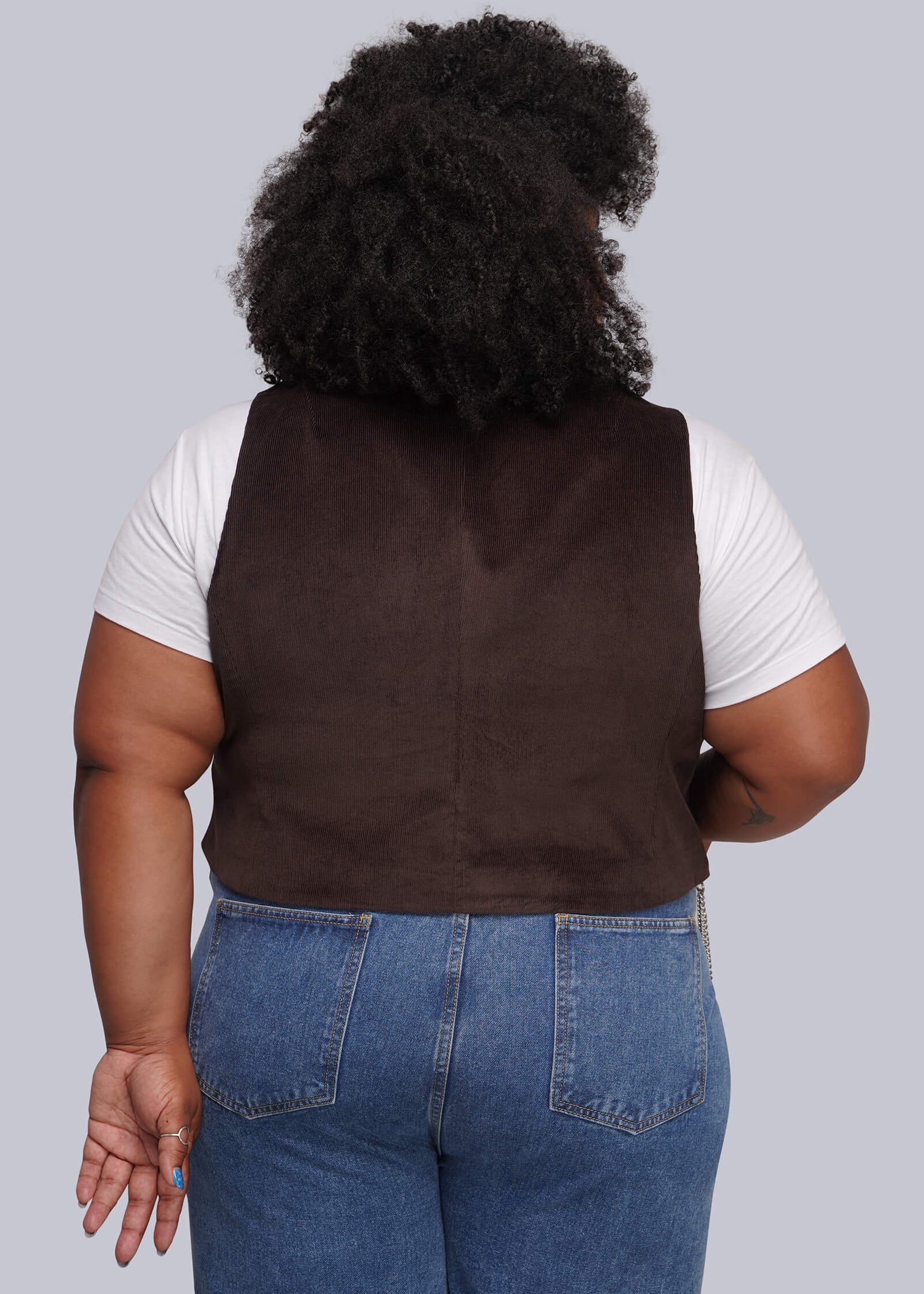 1X=A person shown from the back wearing a dark brown sleeveless corduroy vest over a white short-sleeve shirt and blue jeans, against a plain light gray studio background.