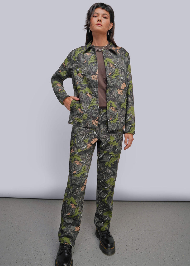 S=A model stands in a studio wearing a matching camouflage jacket and pants set with a brown shirt underneath and black boots. The camouflage pattern features green, brown, and black tones.
