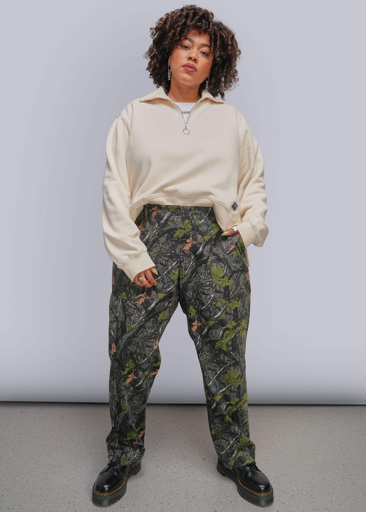 1X=A model stands against a light gray studio backdrop wearing a cream quarter-zip sweatshirt and dark green camouflage pants with black boots.