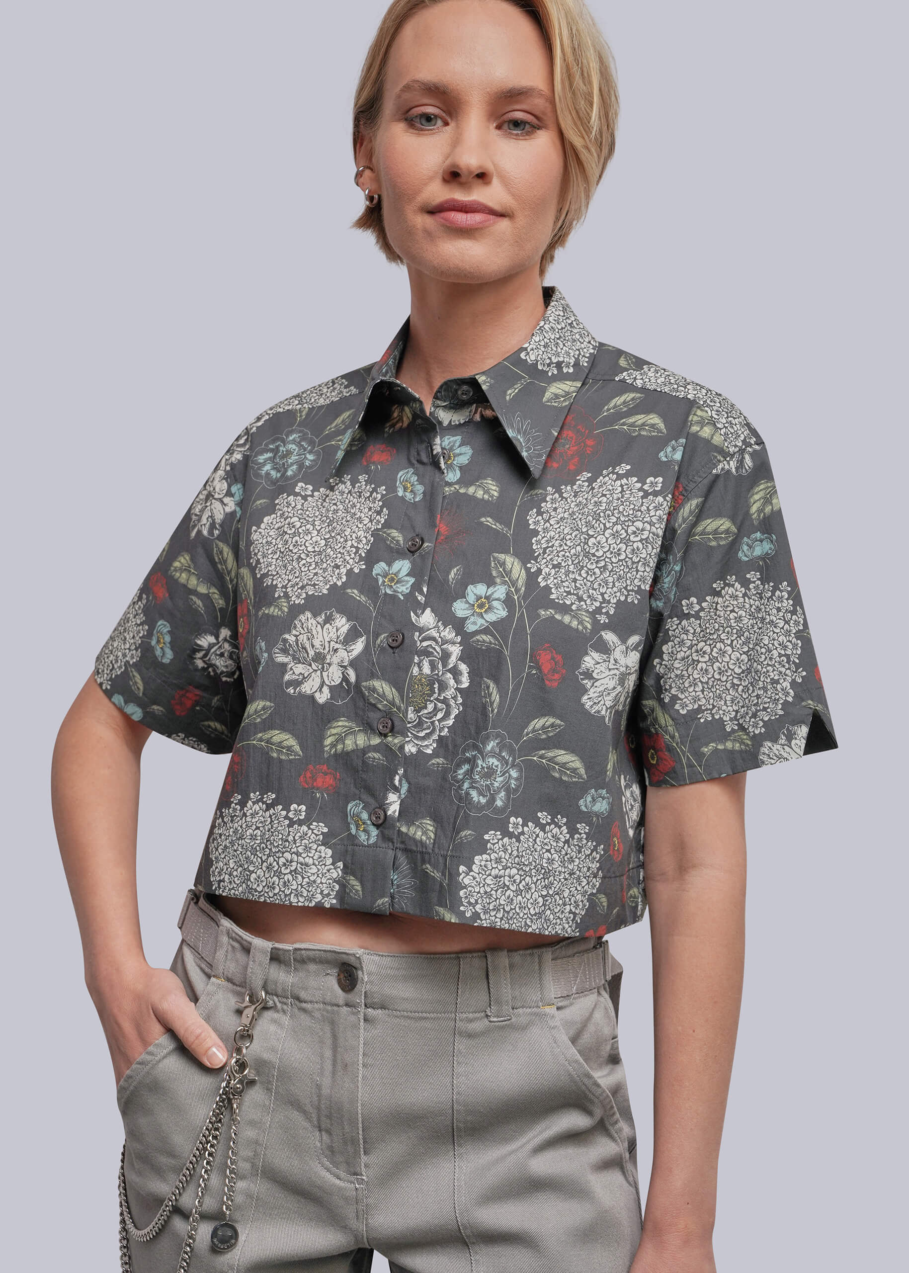 S=A woman wearing a cropped short-sleeve button-up shirt with a dark gray floral print and light gray pants, photographed against a plain light gray studio background.