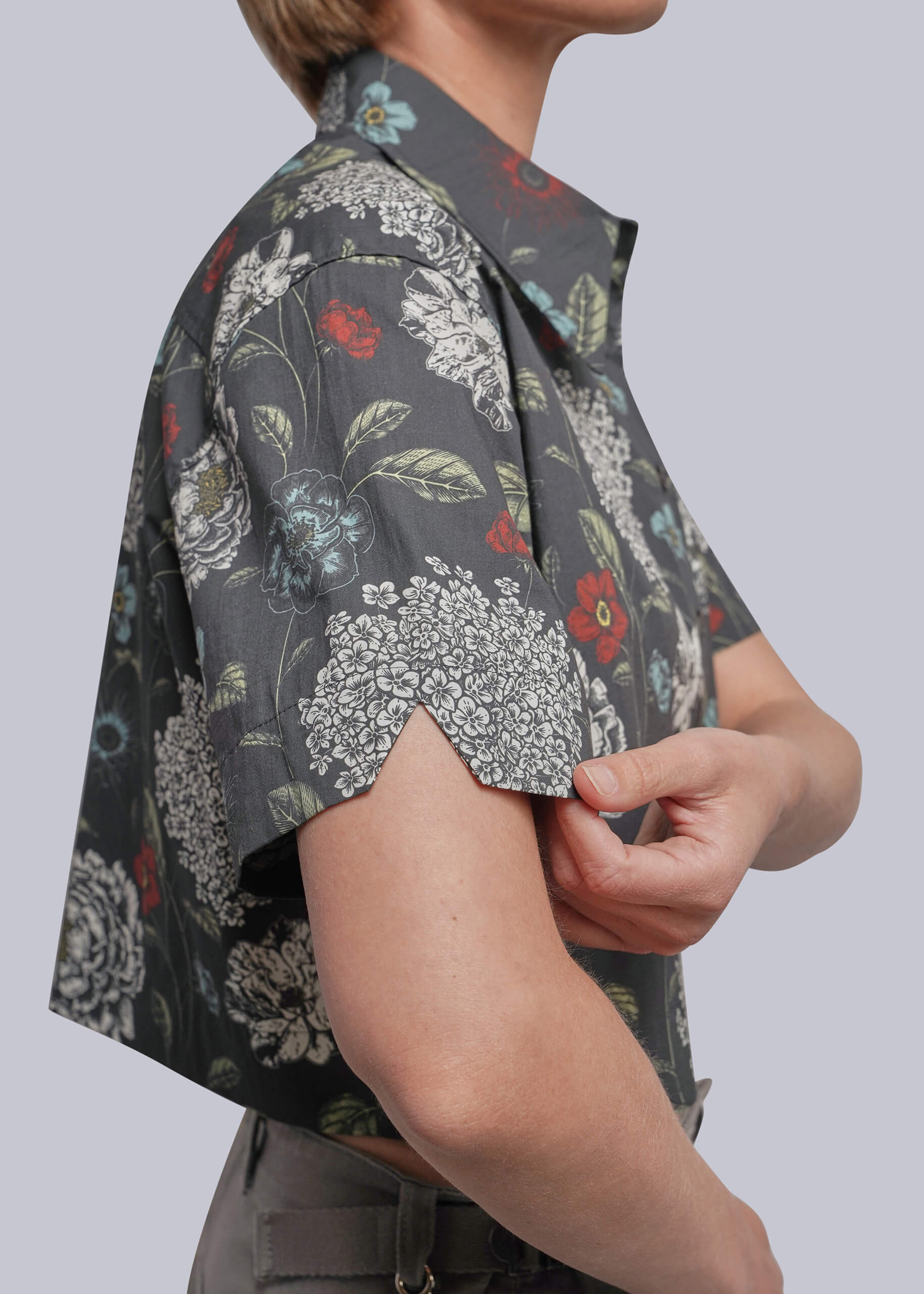 S=A close-up side view of a person wearing a short-sleeve button-up shirt with a dark gray base and a dense white, red, and green floral print. The person is adjusting the cuff of the shirt against a solid light gray background.