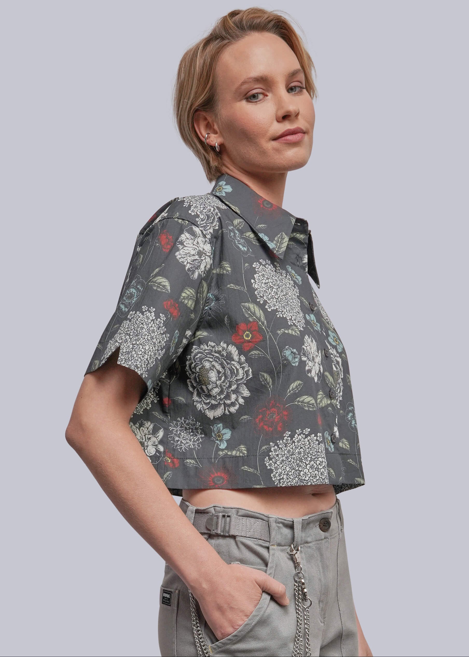 S=A woman wearing a cropped short-sleeve button-up shirt with a gray floral print and light gray trousers, photographed against a solid light gray studio background.