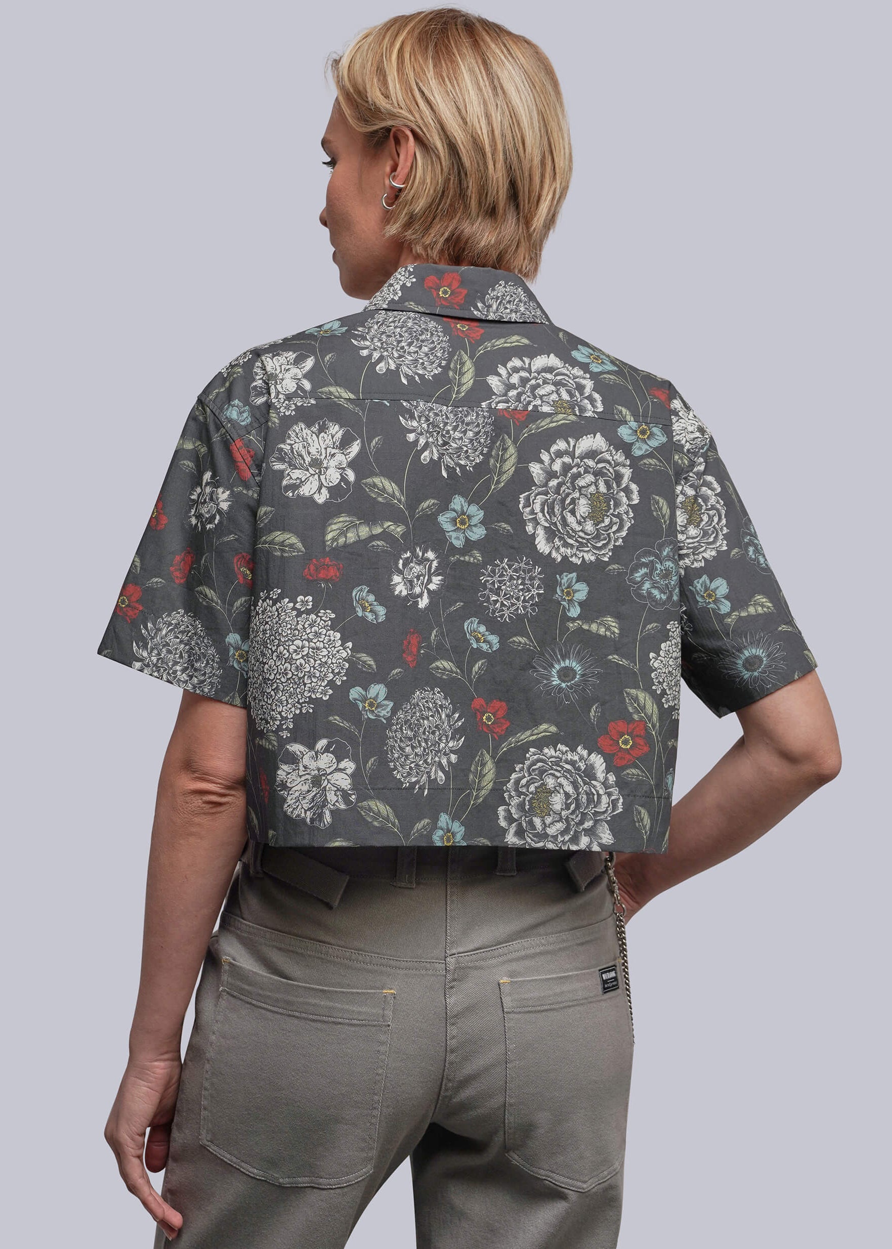 S=A woman shown from the back wearing a short-sleeve button-up shirt with a dark gray base and a white, green, and red floral pattern, paired with olive green pants, against a solid gray background.