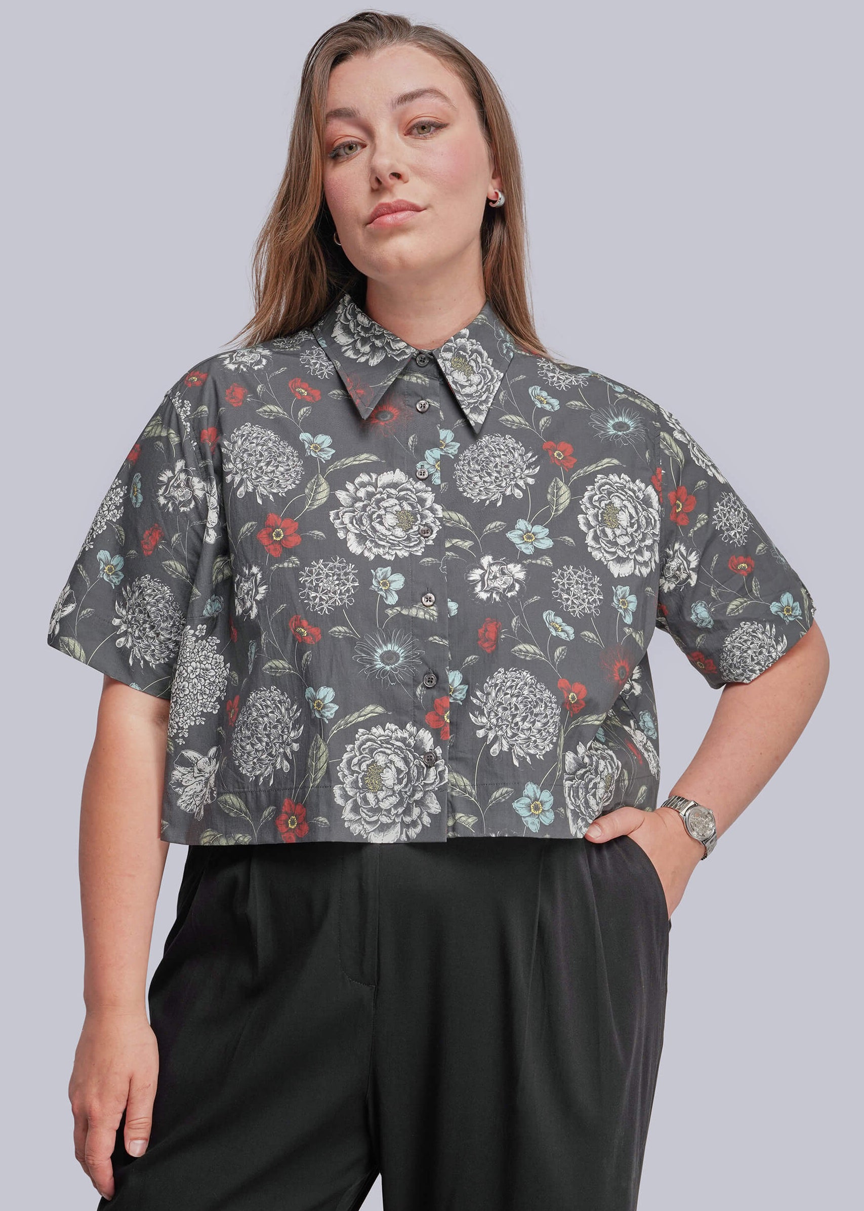 1X=A woman modeling a short-sleeve button-up shirt with a gray background and a white, red, and blue floral pattern, paired with black trousers, against a solid gray studio background.