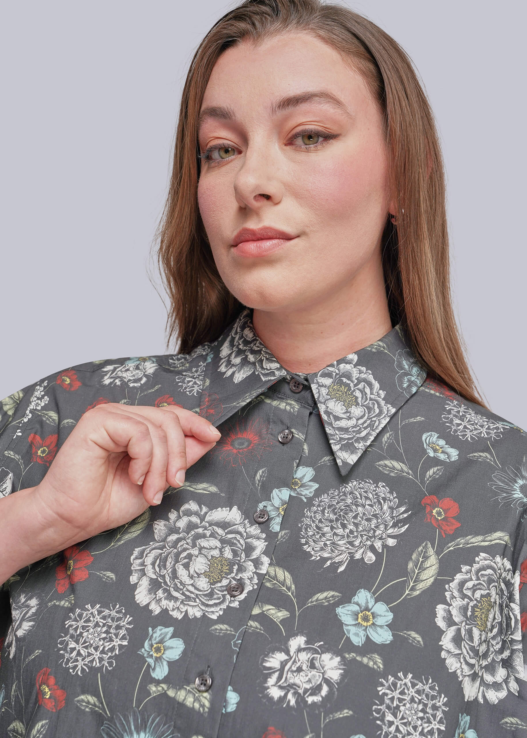1X=A close-up of a woman wearing a gray button-up shirt with a detailed floral print in white, light blue, and red, set against a solid light gray background.