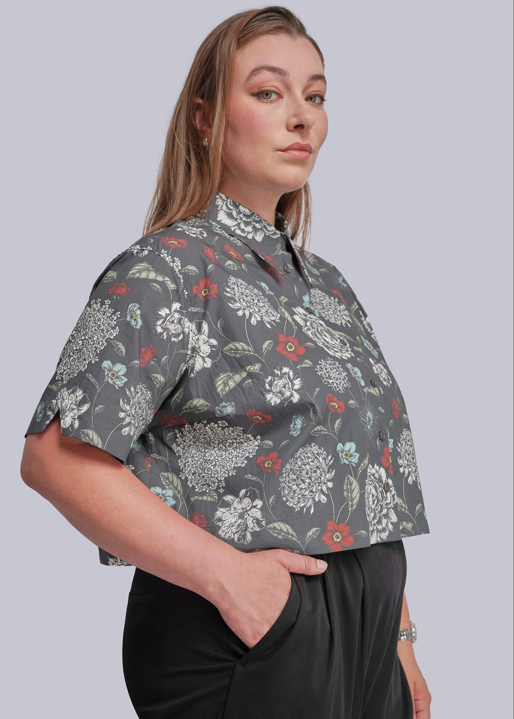1X=A woman wearing a short-sleeve gray button-up shirt with a white, red, and green floral pattern, paired with black trousers, against a solid gray background.