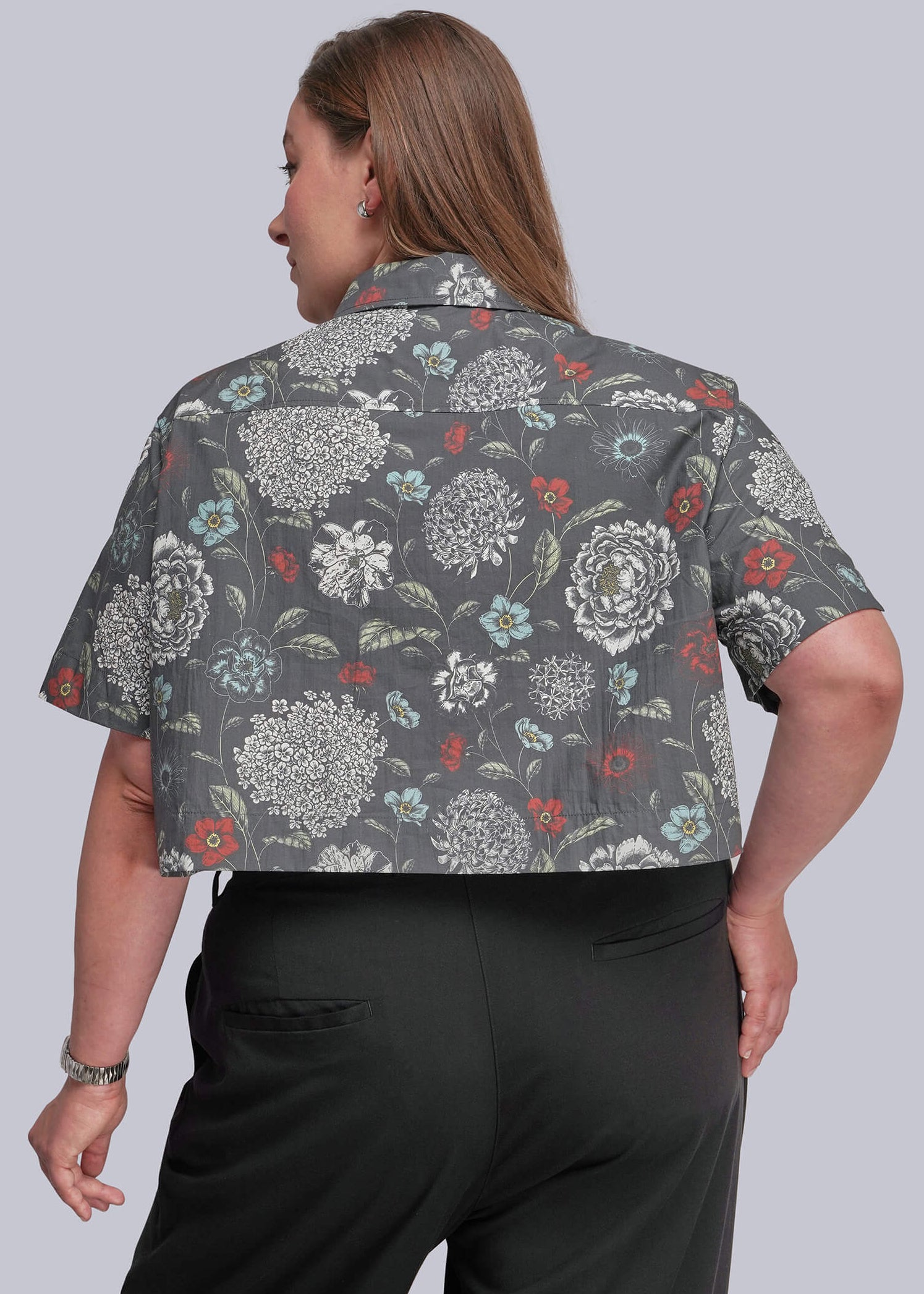 1X=A woman shown from the back wearing a short-sleeve button-up shirt with a gray floral pattern and black trousers, against a solid light gray background.