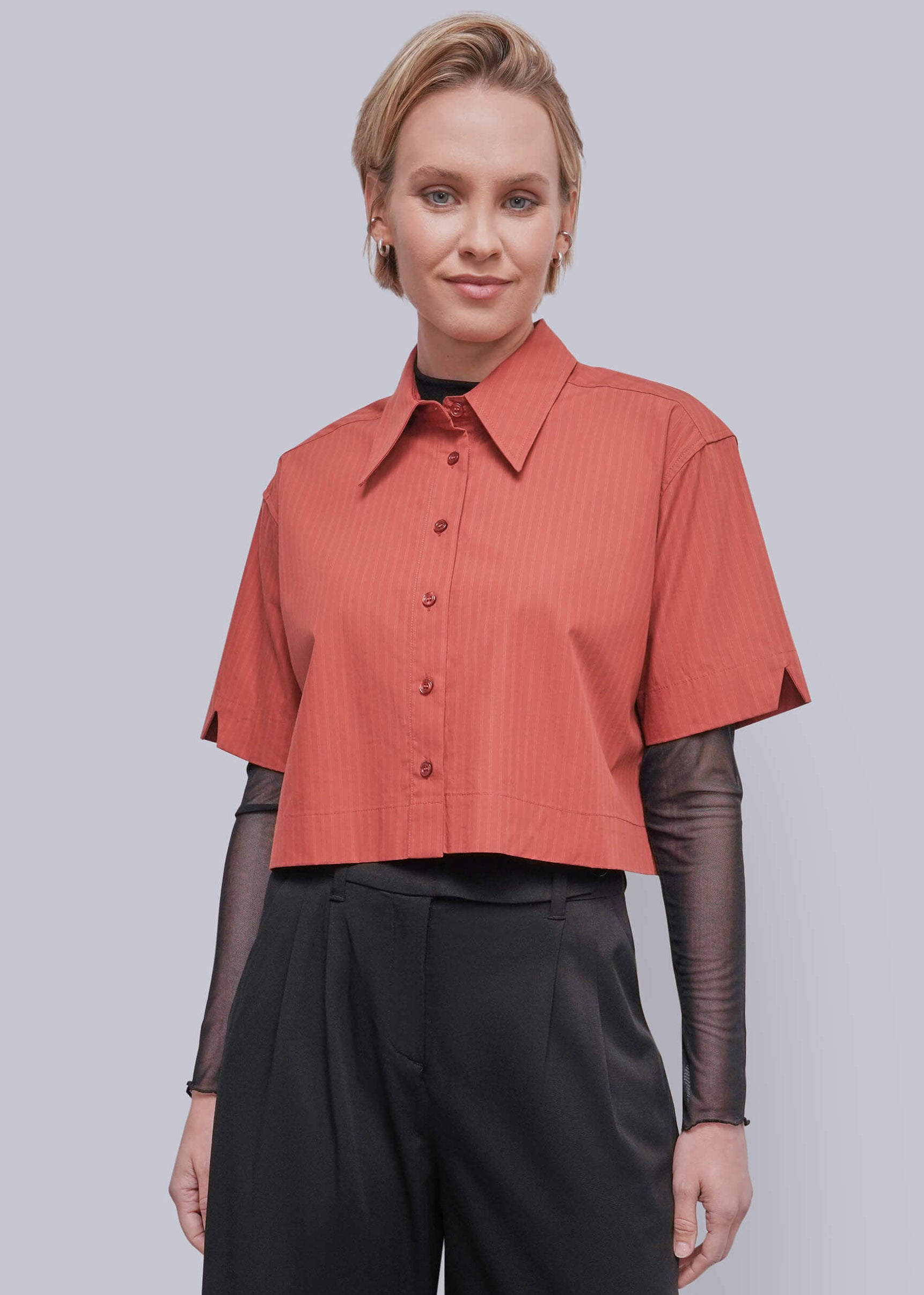 S=A woman modeling a short-sleeve cropped button-up shirt in a muted terracotta or burnt orange color with subtle vertical texture, layered over sheer black long-sleeve sleeves and black trousers, against a solid light gray studio background.