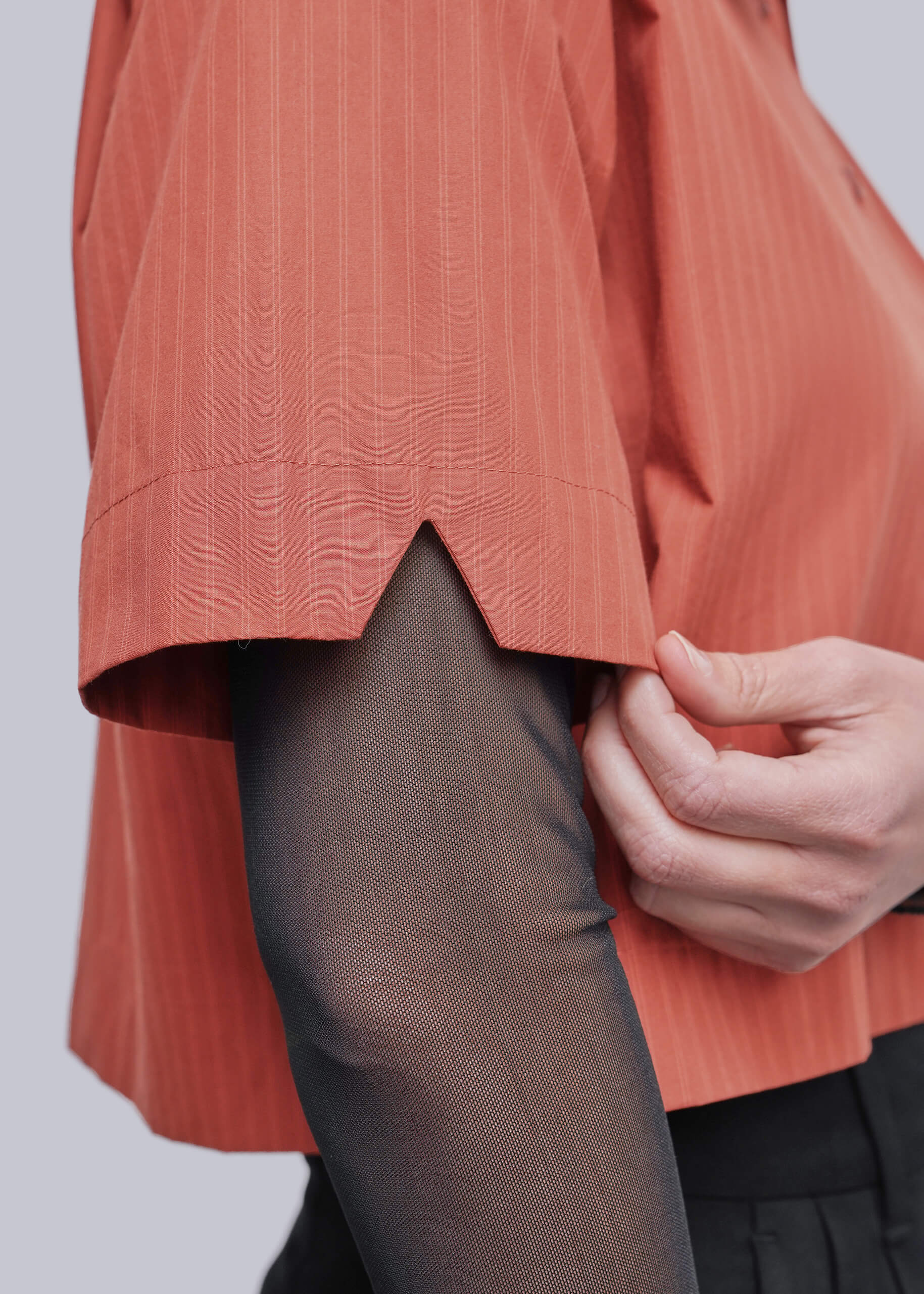 S=Close-up of a person wearing a short-sleeve terracotta orange pinstriped shirt with a hand adjusting the hem, paired with sheer black tights and dark trousers, against a light gray background.