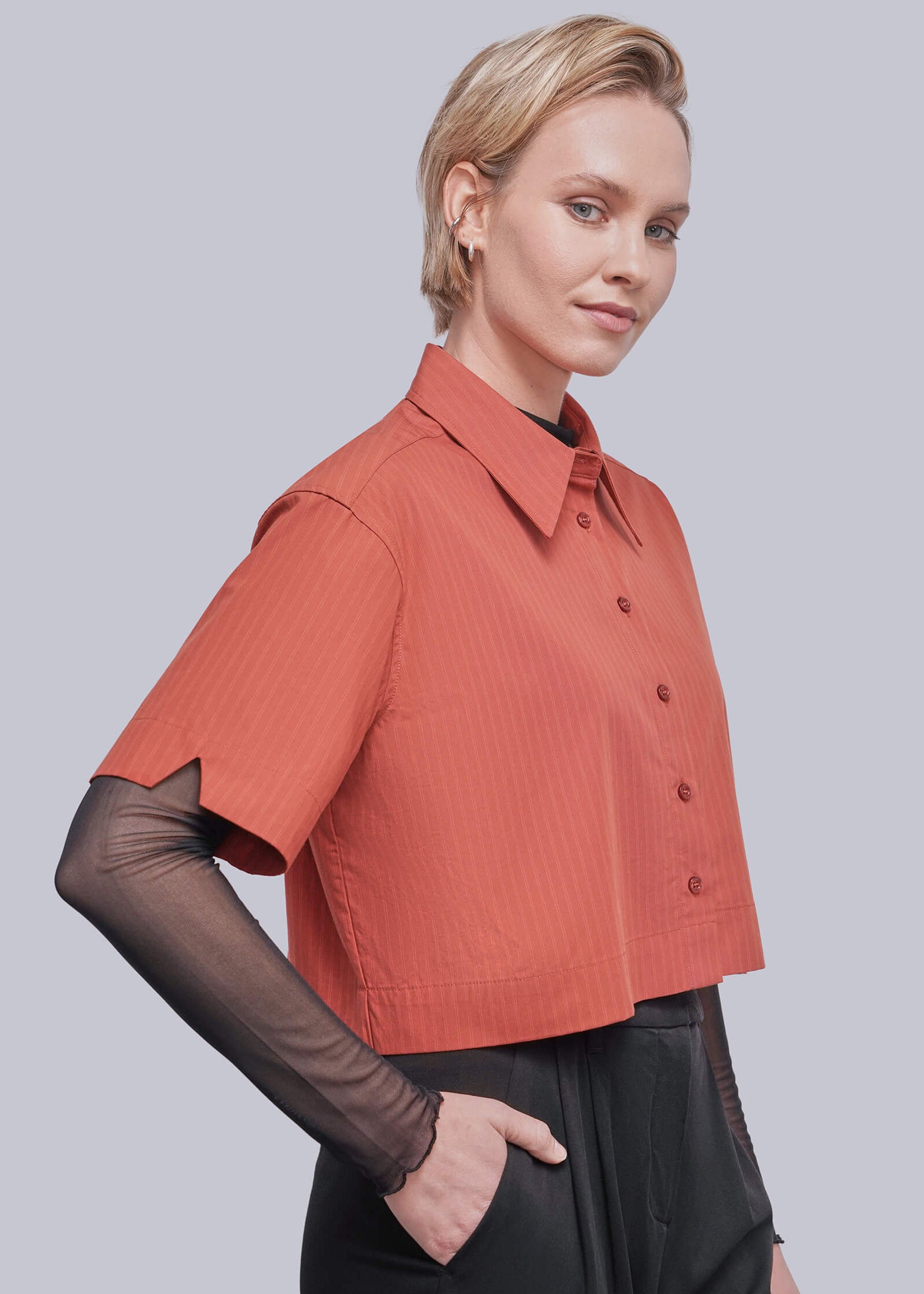 S=A woman modeling a short-sleeve collared button-up shirt in a muted terracotta or burnt orange color, layered over sheer black long sleeves, paired with black trousers, against a solid gray studio background.