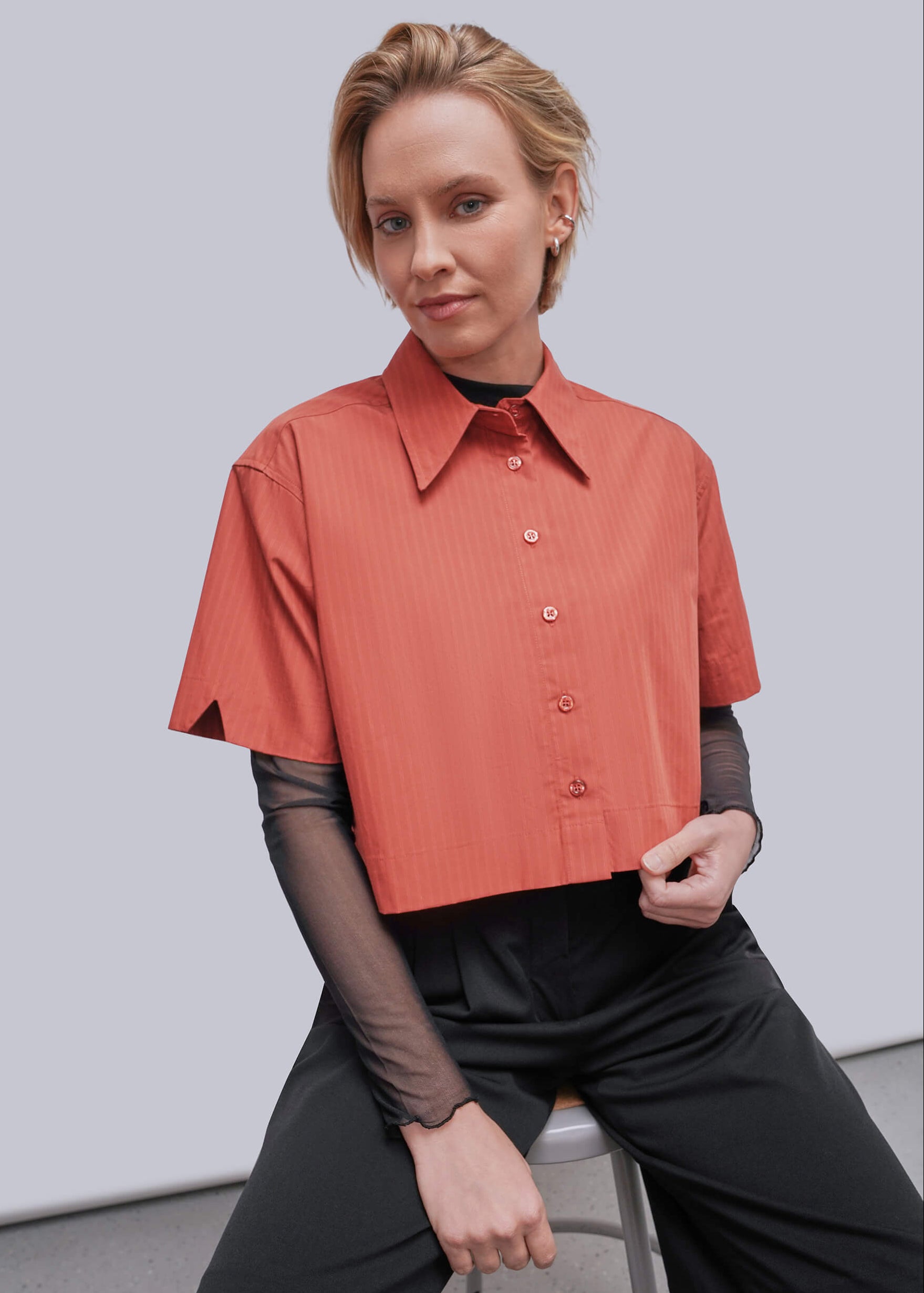 S=A woman seated on a stool wearing a short-sleeve collared button-front top in a muted coral-orange tone over sheer black long-sleeve sleeves, paired with black pants, against a plain light gray studio background.