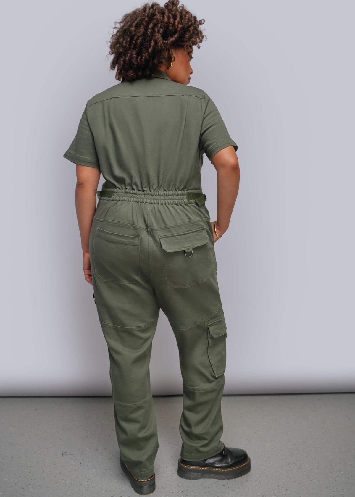 1X=A person shown from the back wearing an olive green short-sleeve utility jumpsuit with cargo pockets, standing against a plain light gray studio background.