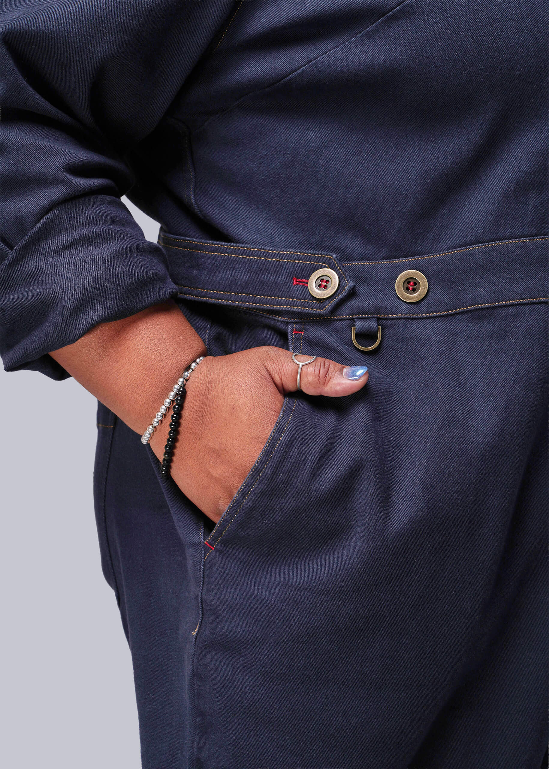 1X=Navy blue jumpsuit with buttons and a person's hand in the pocket on a light gray background