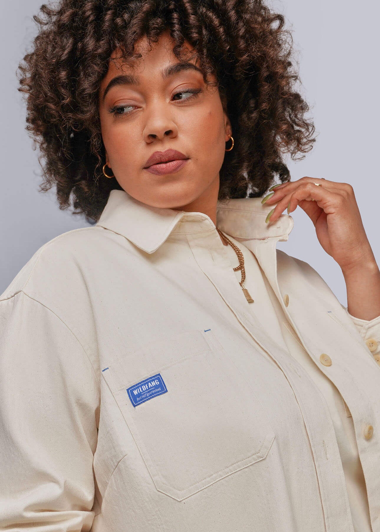 1X=A close-up of a woman wearing a light beige button-up shirt jacket with a blue 'WILDFANG' brand patch on the chest pocket, set against a plain light gray background.