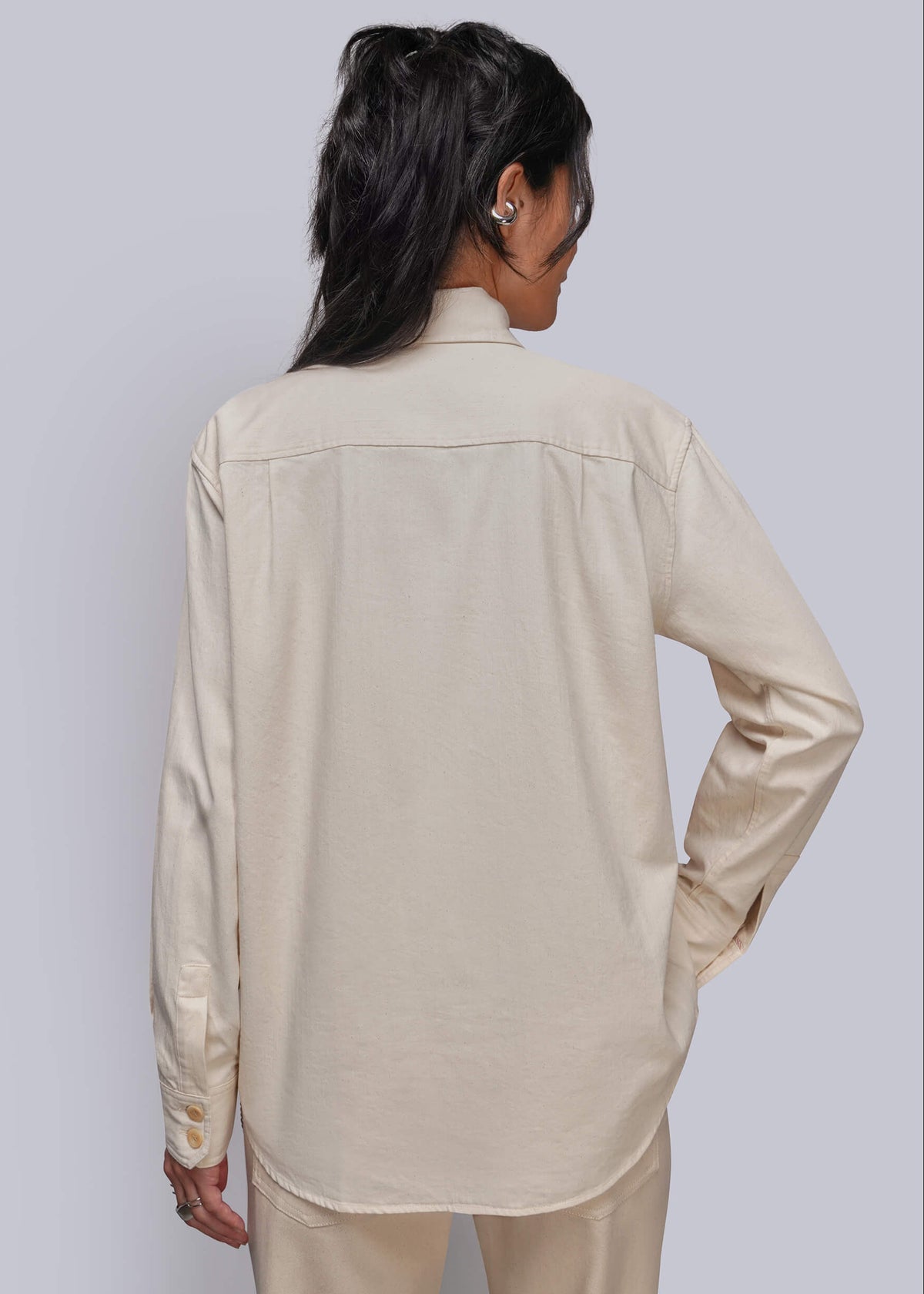 S=A woman shown from the back wearing a light beige long-sleeve button-up shirt and matching beige pants against a light gray studio background.