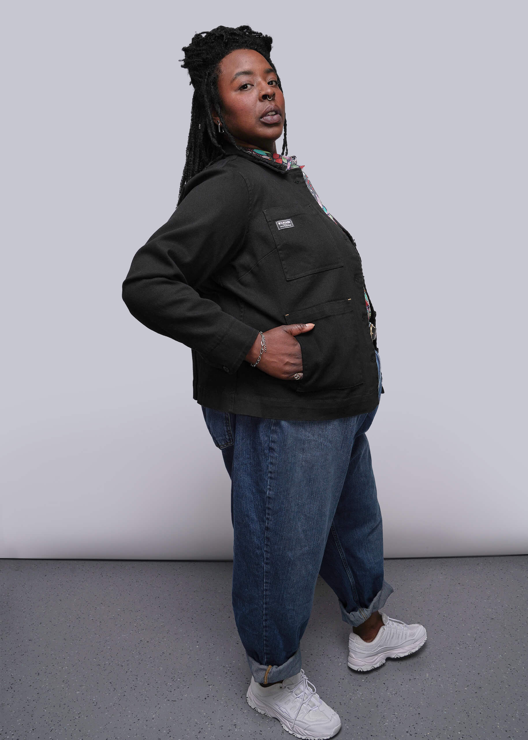 1X=Person wearing a black denim jacket, blue jeans, and white sneakers against a plain background