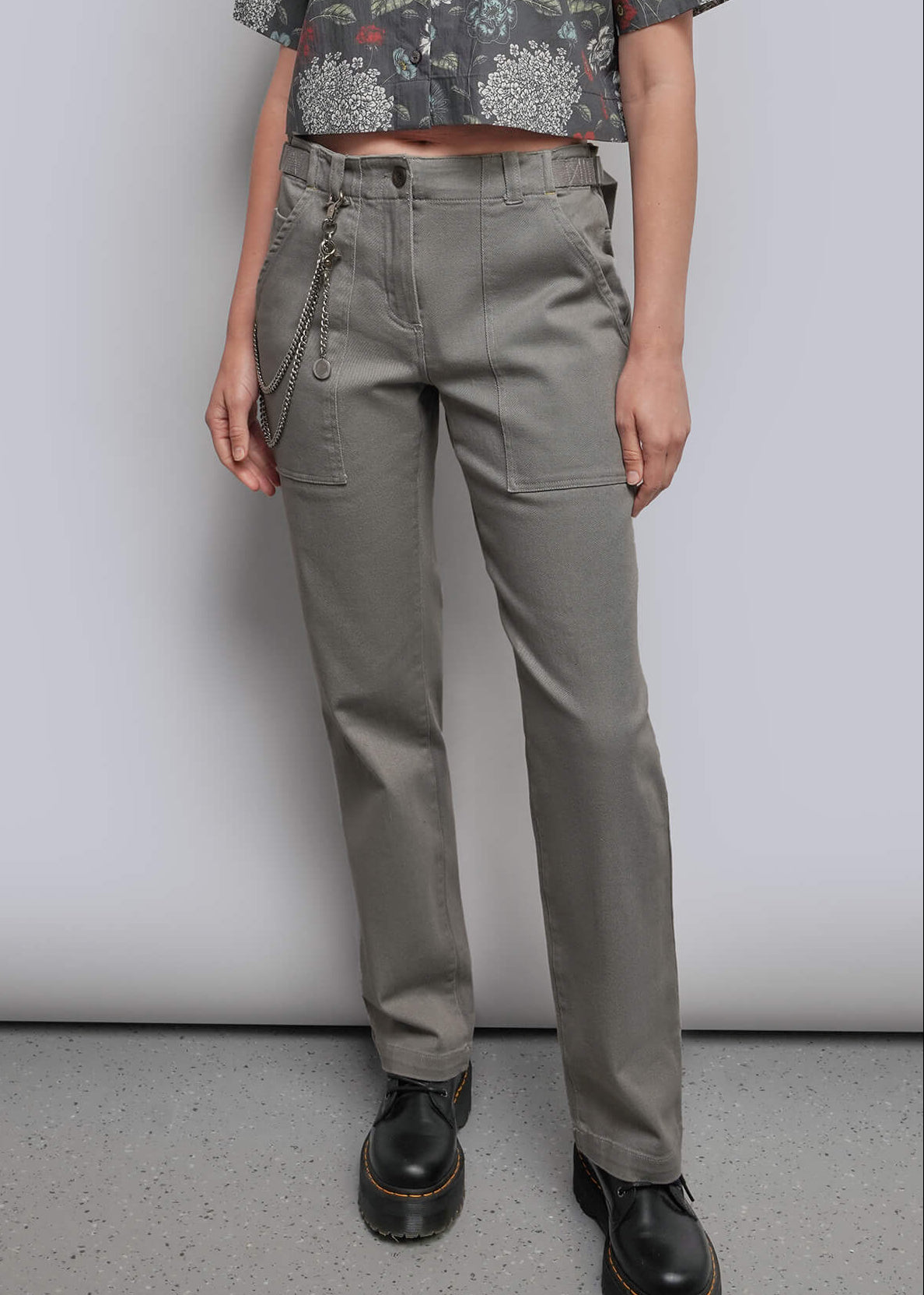 S=A model stands against a light gray studio background wearing a short-sleeve button-up shirt with a muted floral print in gray, green, and burgundy, paired with olive-gray trousers and black shoes.