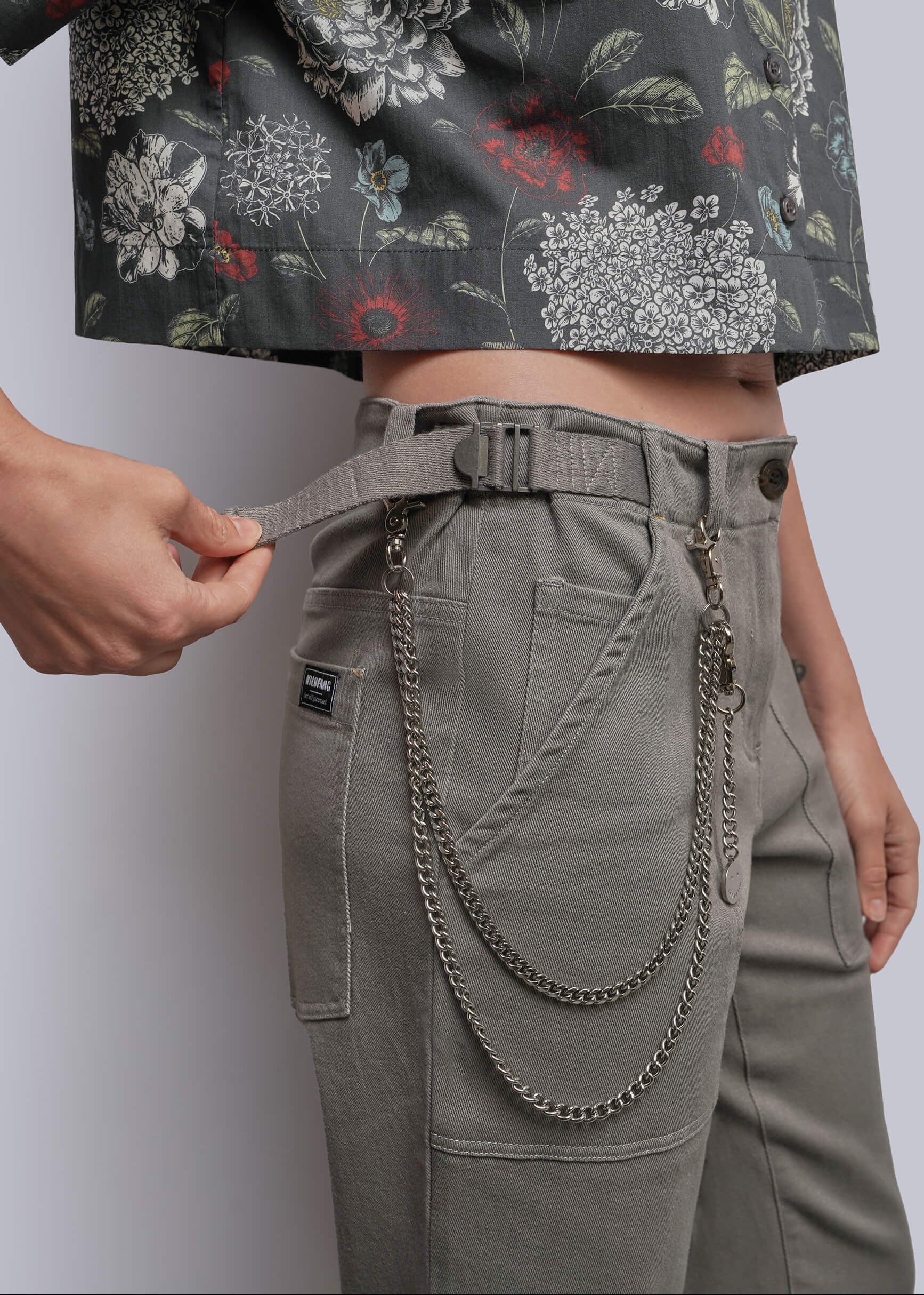 S=Close-up of a person wearing high-waisted gray cargo-style pants with a matching belt and hanging metal chain detail, paired with a cropped floral top against a light gray studio background.