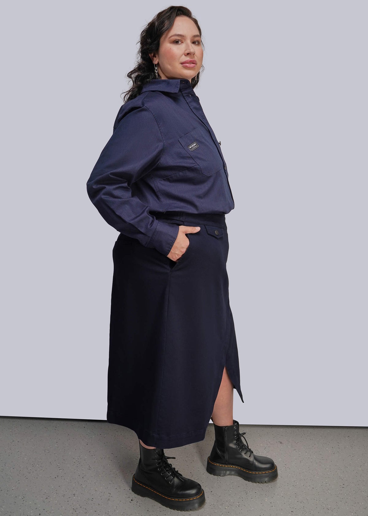 1X=A woman modeling a navy blue button up and matching wrap skirt set, paired with black combat boots, against a light gray studio background.