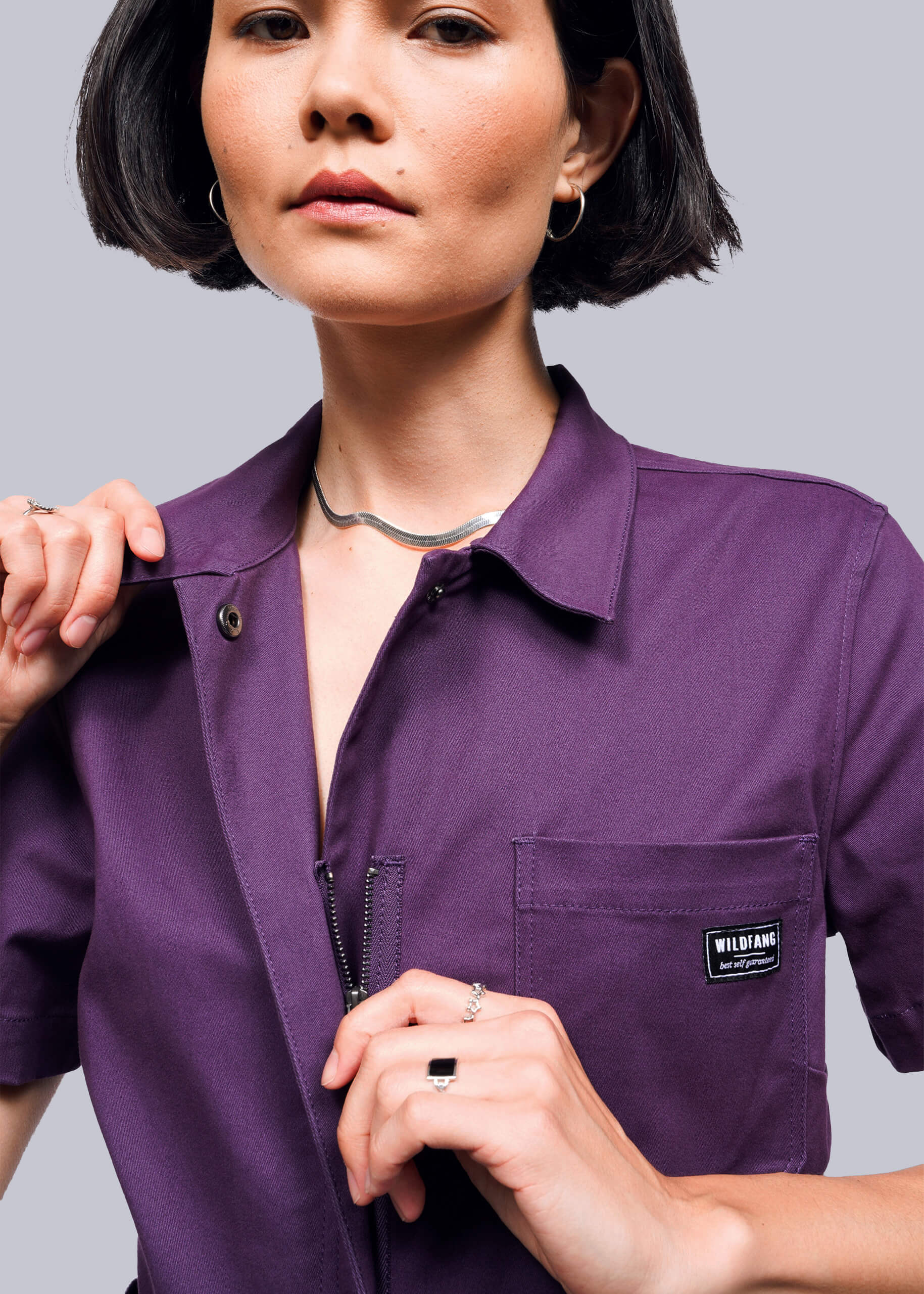 S=Close up of person wearing a purple jumpsuit with a brand logo on a gray background
