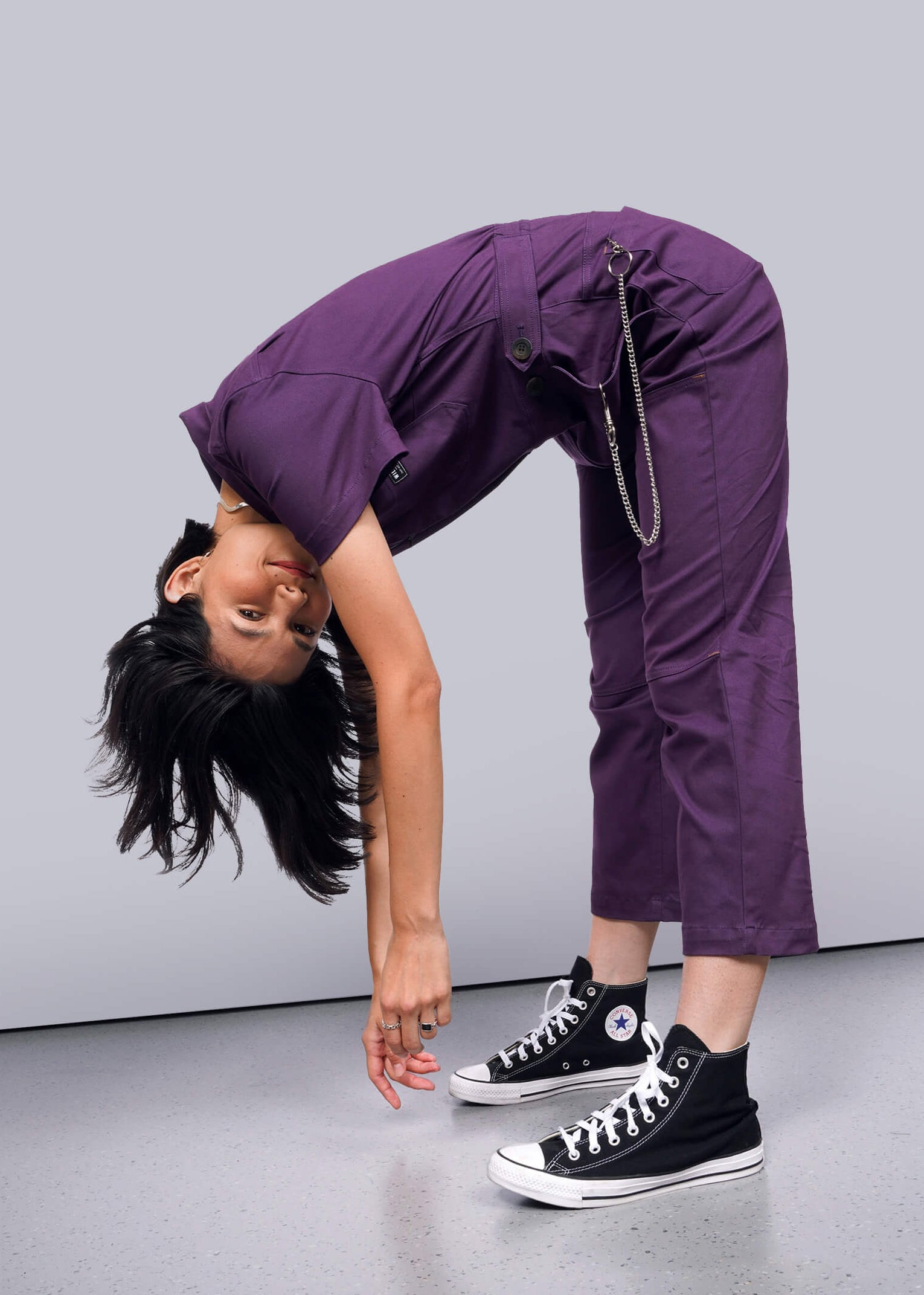 S=Person wearing a purple jumpsuit with black high-top sneakers on a gray background